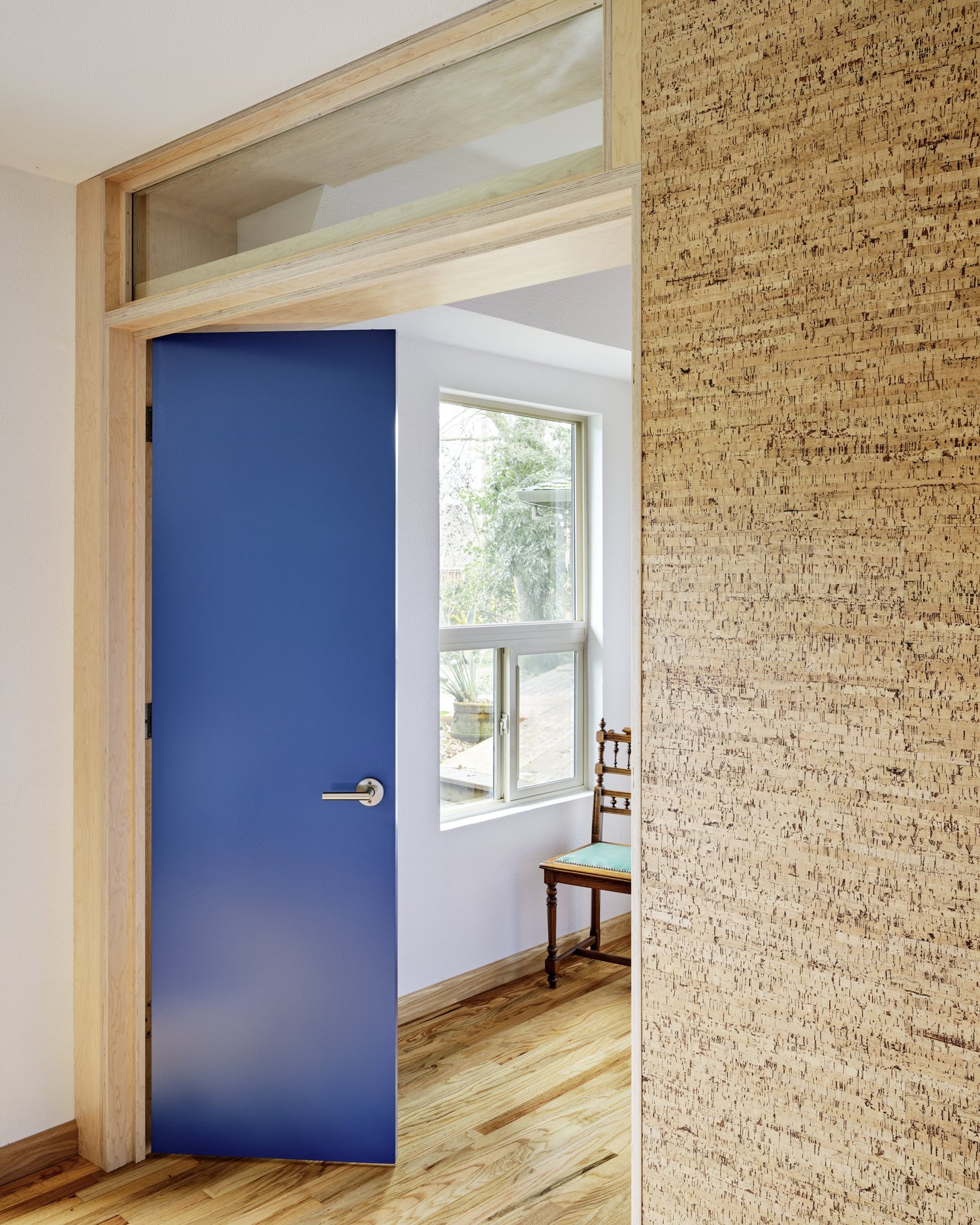 Interior of Three Cork Walls showing a vivid blue door opening to a bedroom with a wooden chair and large window, framed by cork wall panels.