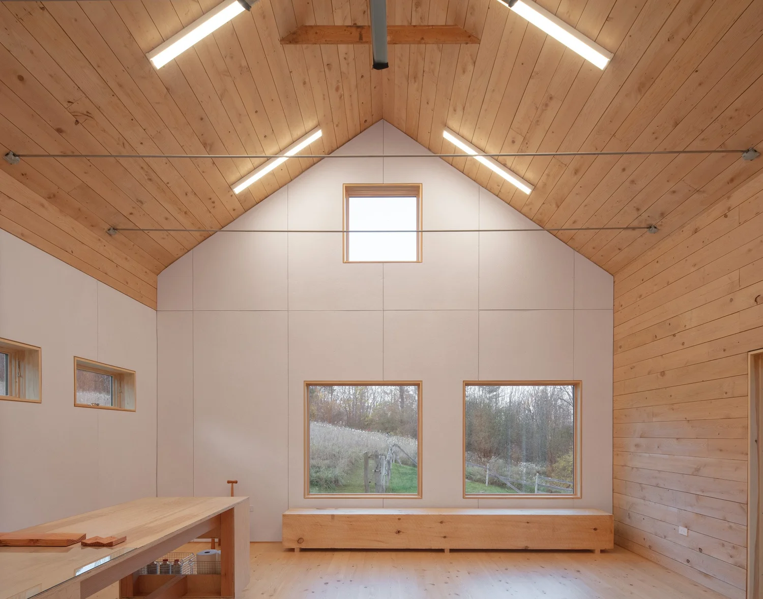 Interior view of Barnard Barn with a pitched wooden ceiling, exposed beams, and rectangular windows framing views of the surrounding landscape. A long wooden bench sits below the windows, and a simple wooden work table anchors the space.
