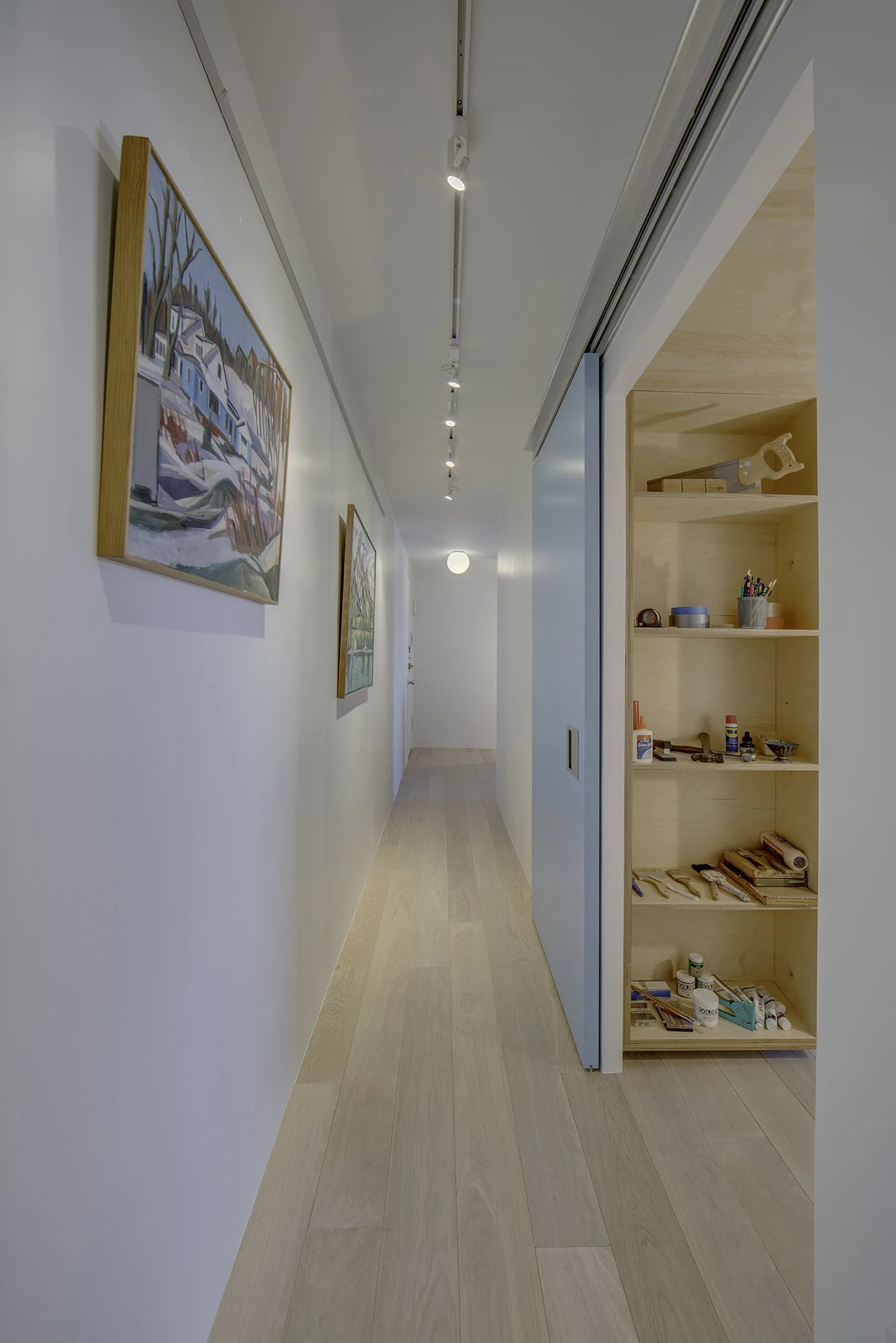 Long hallway at Harlem Studio with light wood floors, white walls, track lighting, and a blue sliding door opening to shelving. Artwork adds color to the minimal design.