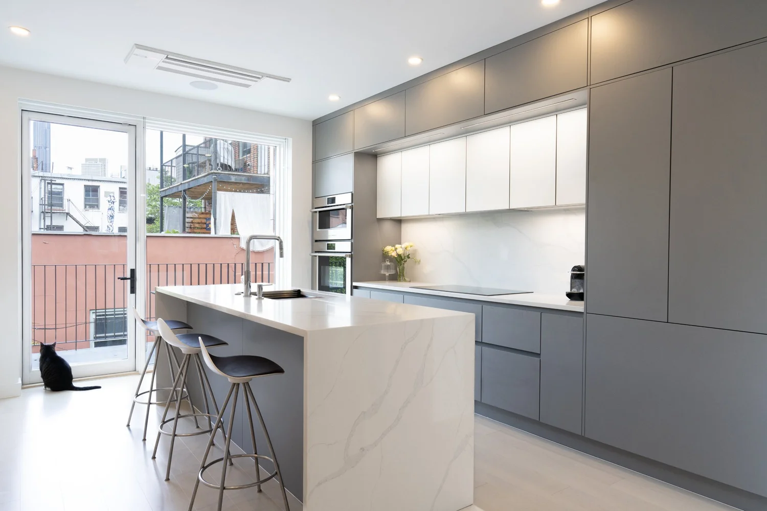 Modern kitchen at Narrow Townhouse with marble island, gray cabinetry, and glass doors opening to a terrace, with a cat sitting by the window.