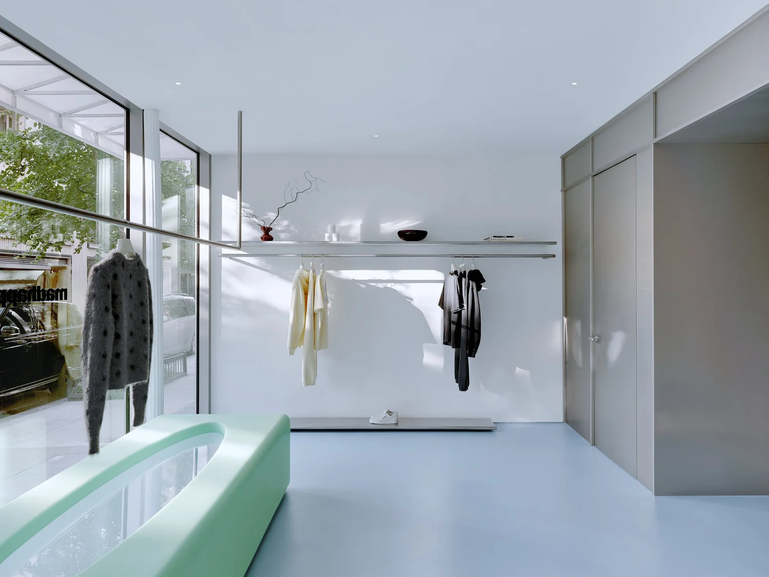 Retail display area inside Madhappy New York Flagship, featuring minimalist clothing racks, a sculptural mint-green bench, and floor-to-ceiling windows.