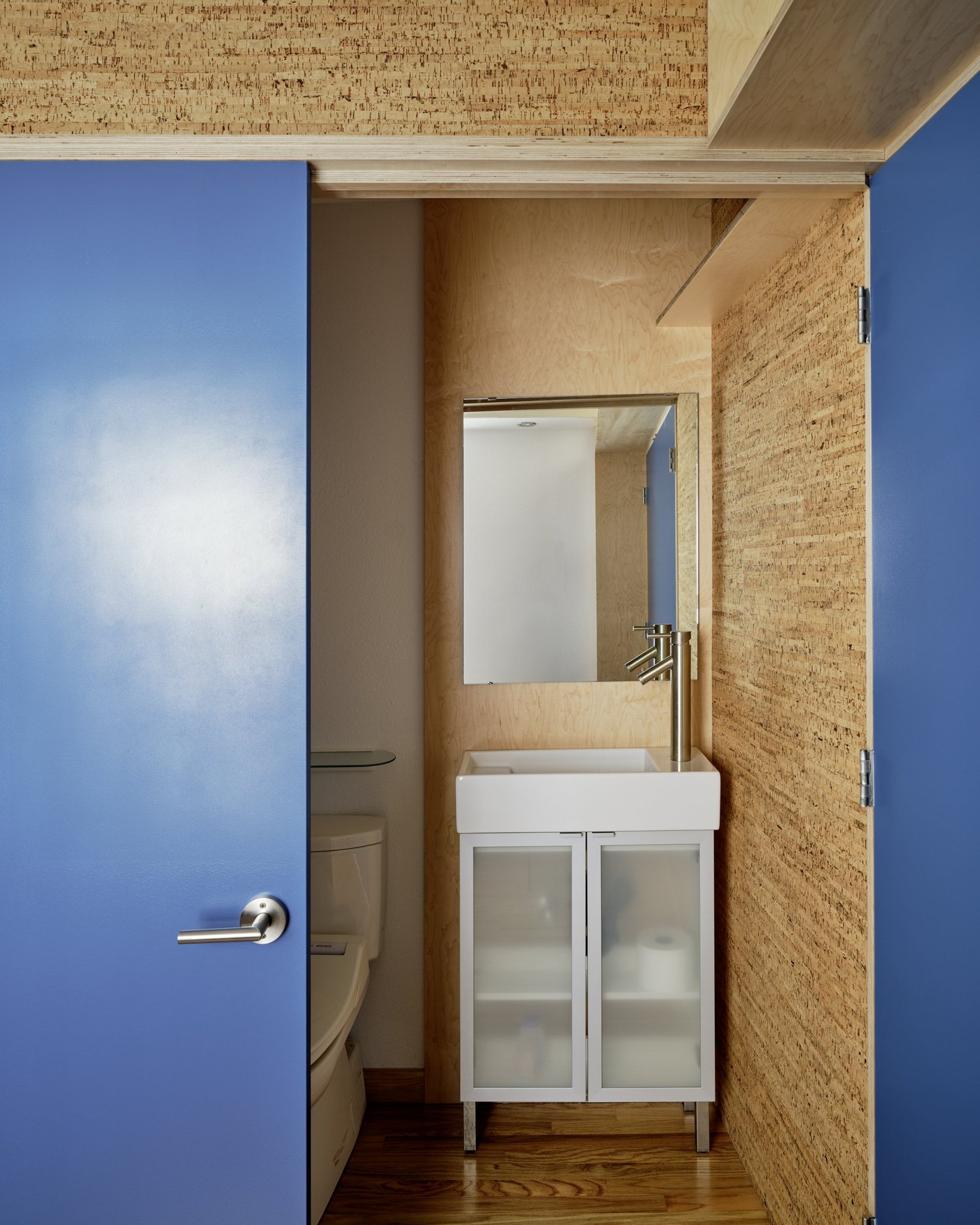 Small bathroom at Three Cork Walls with cork-clad walls, a blue door, white sink cabinet, and mirror above.