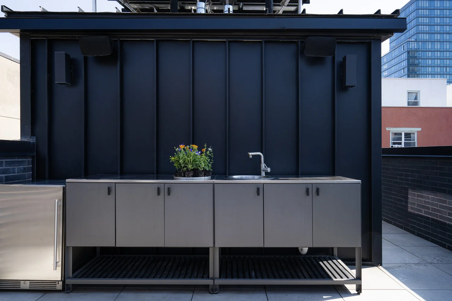 Rooftop kitchen at Narrow Townhouse featuring matte black wall cladding, stainless steel cabinetry, sink, and planter with flowers.