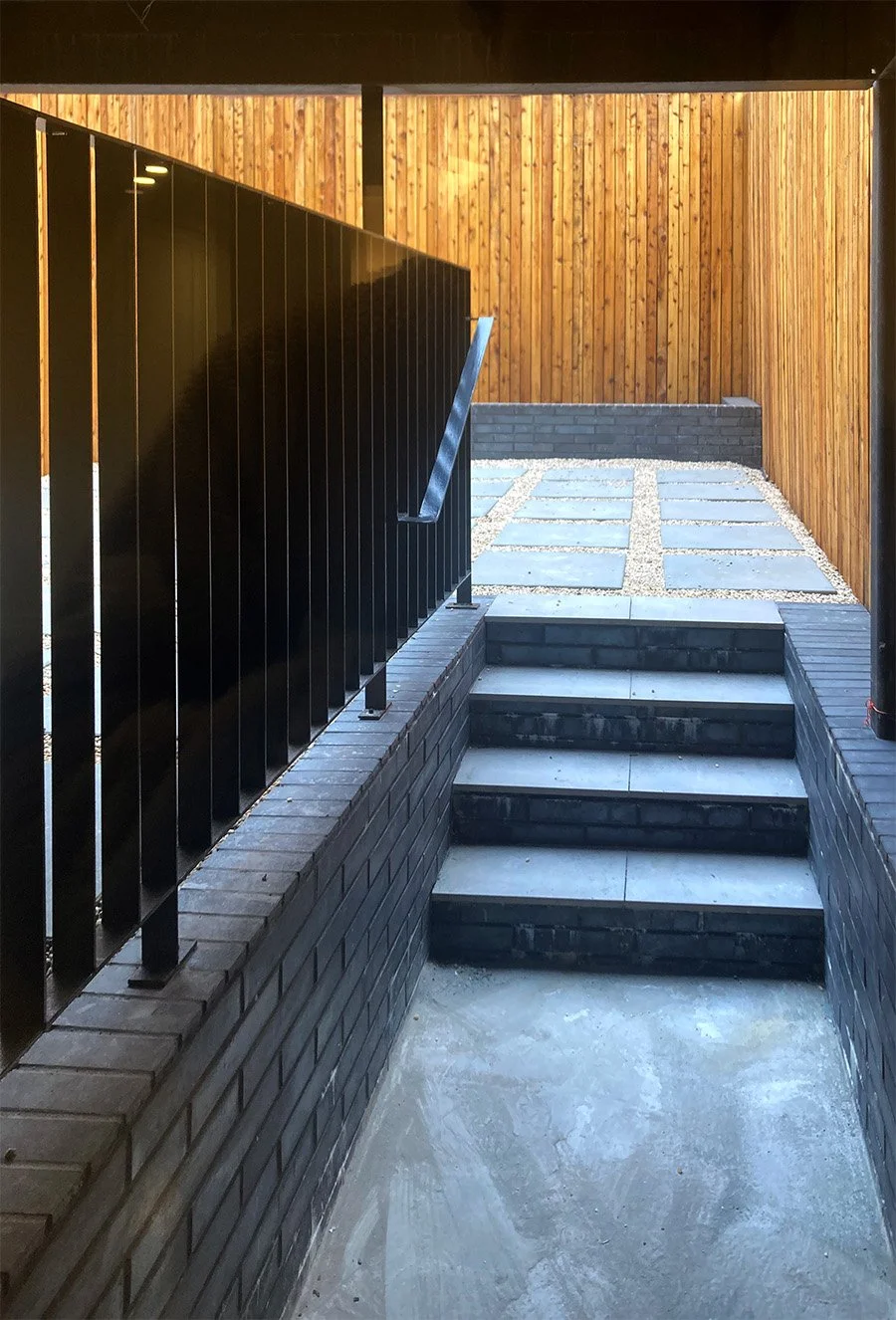 Stairway with dark brick walls and metal railing leading up to a terrace with wood fencing.