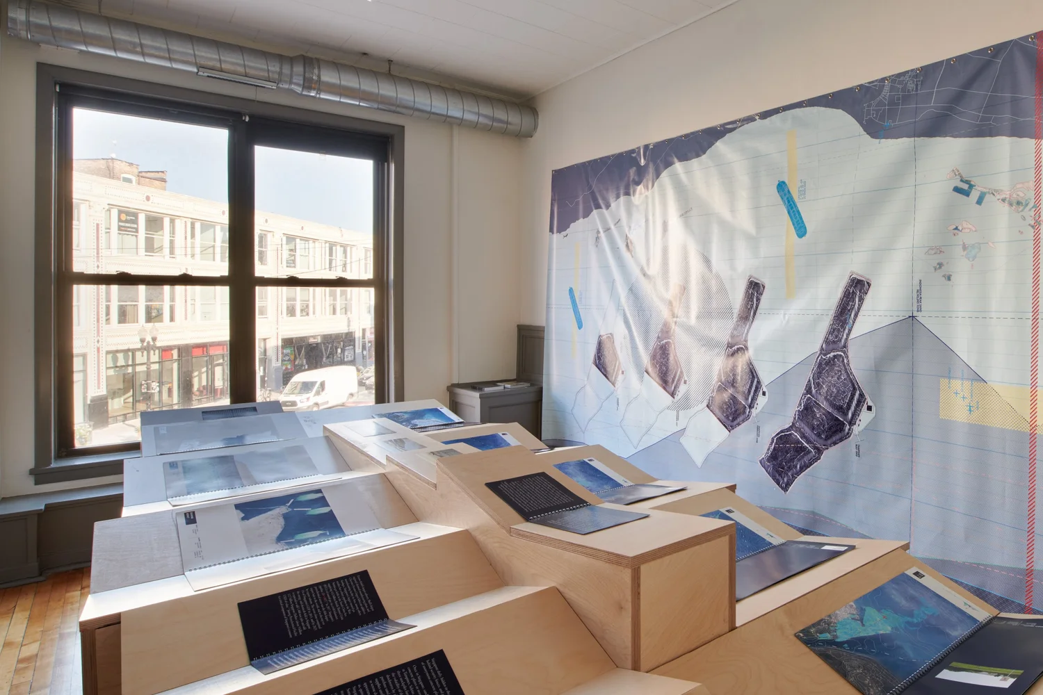 Exhibition view of Territories of Territory Extraction showing a stepped wood display with open books and research materials, alongside a large wall map, in a gallery with street-facing windows.
