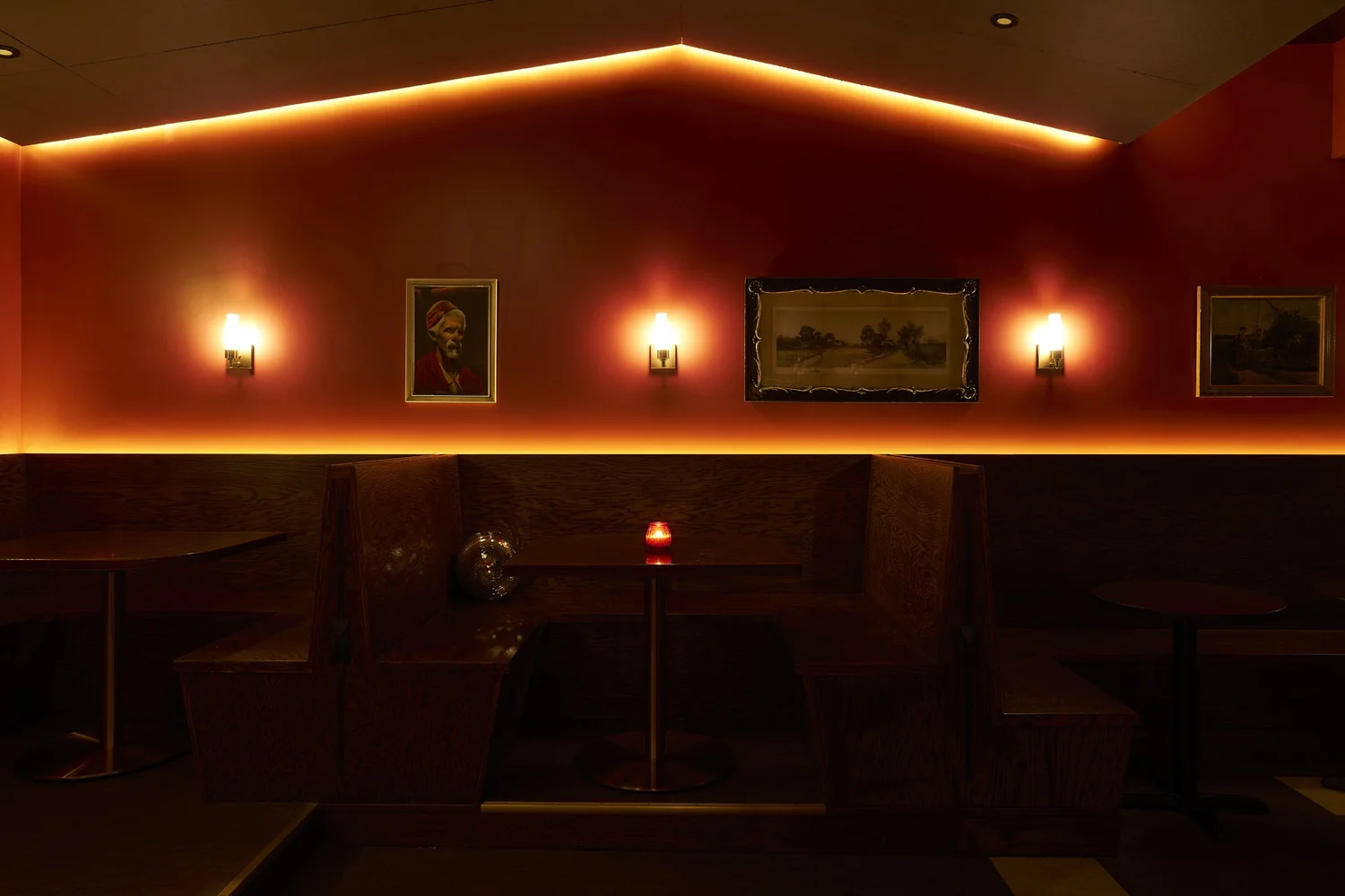 Booth seating at Mr. Nancy’s with dark wood benches, red-painted walls, wall sconces, and framed artwork illuminated by warm accent lighting.