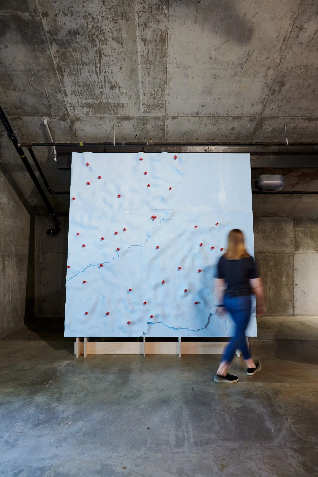 Installation view of The Great Lakes Architectural Expedition showing a large blue relief map with red markers, mounted against a concrete wall, with a person walking past.