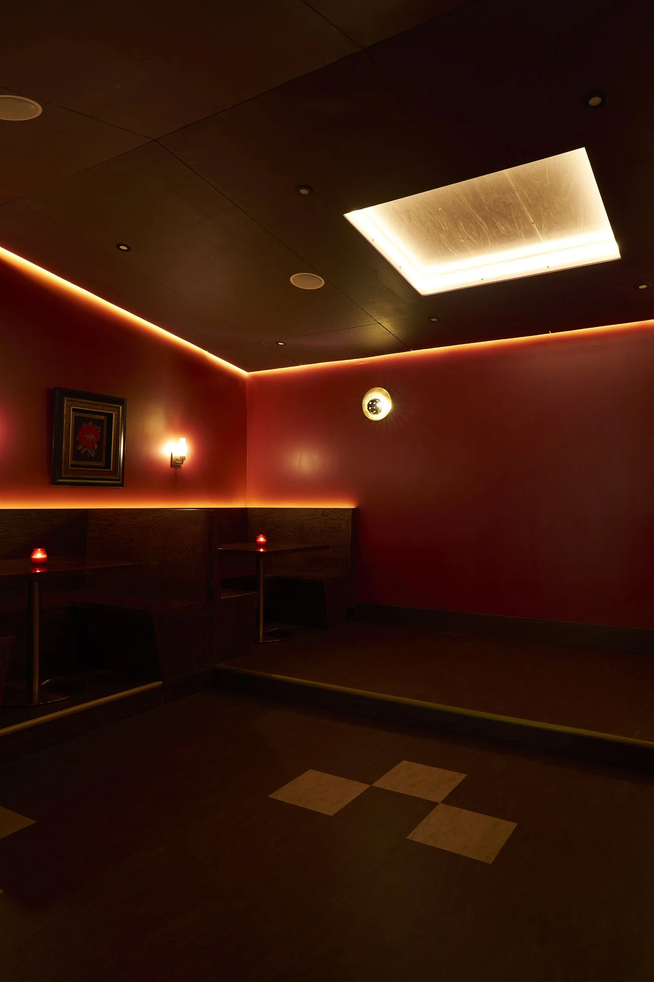 Lounge area at Mr. Nancy’s with red-painted walls, glowing perimeter lighting, small tables, and framed artwork illuminated by wall sconces.
