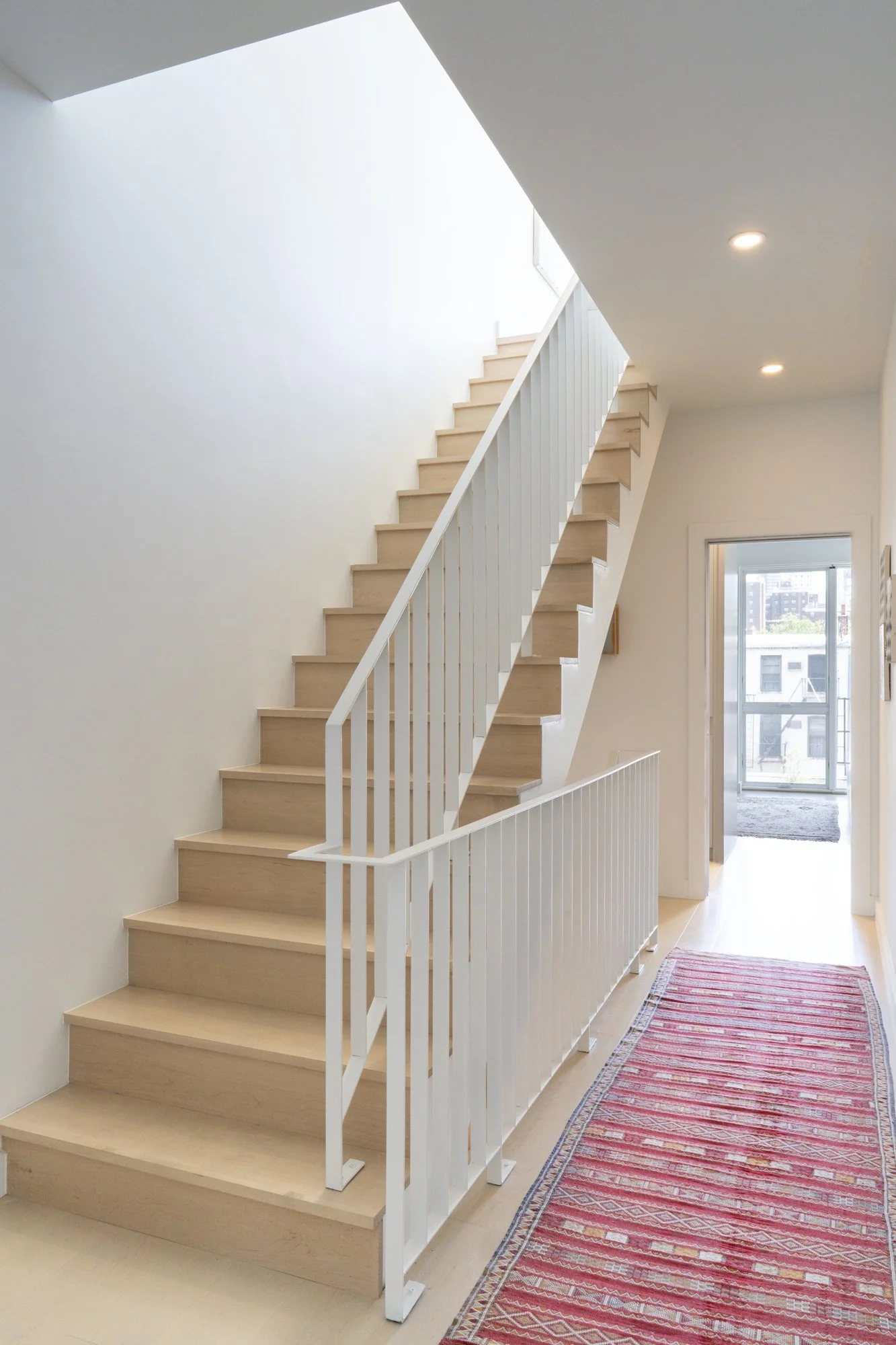 Interior view of Narrow Townhouse central staircase with light wood treads, white railing, and natural daylight from above.