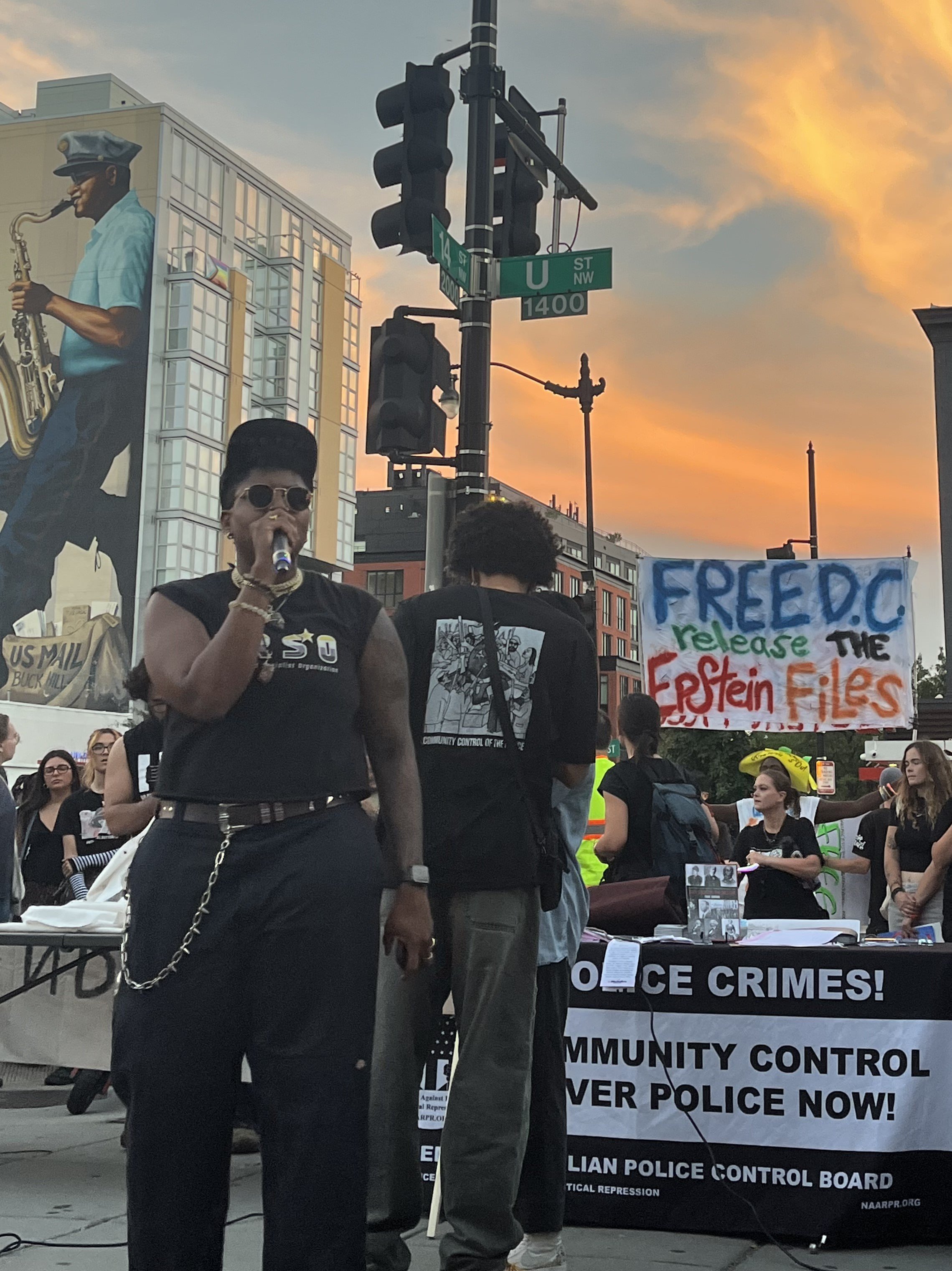 Protest scene in an urban area at sunset, featuring a person speaking into a microphone, signs advocating for community control of police and releasing Epstein Files, alongside a large mural of a man playing the saxophone.