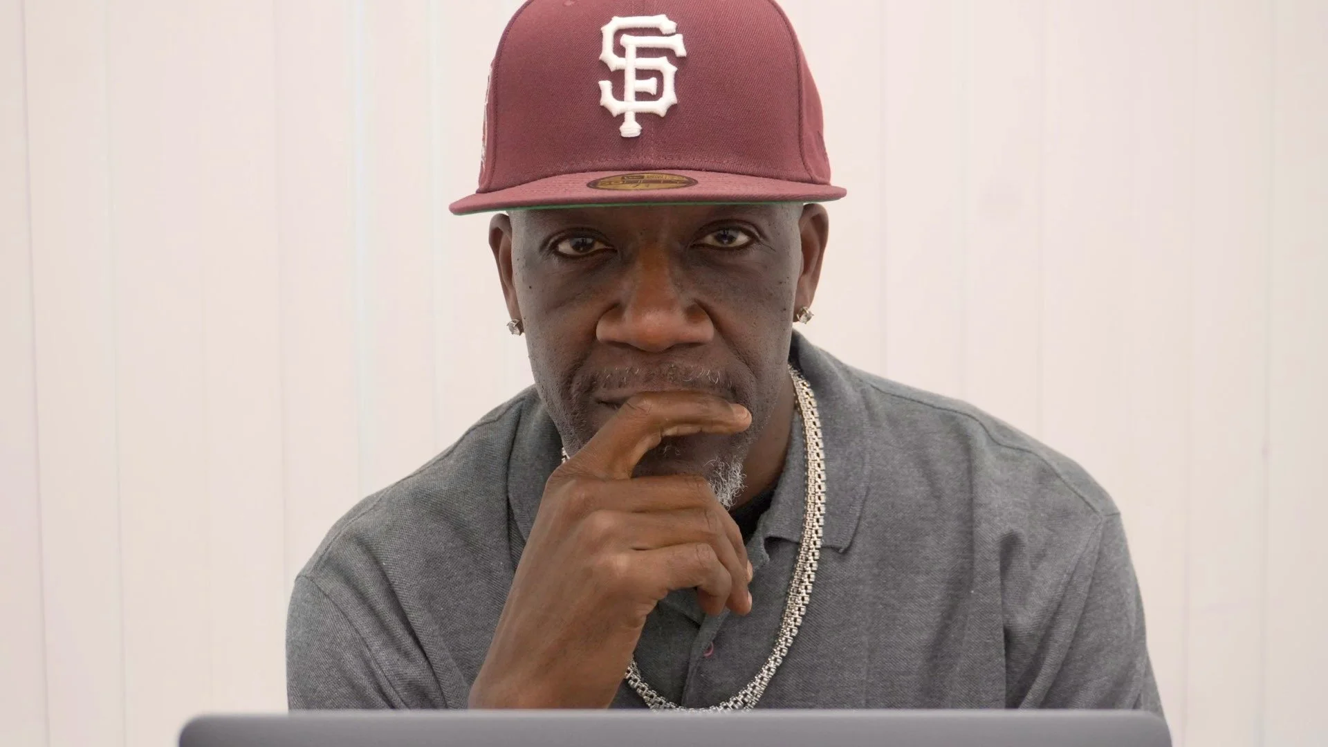 A man wearing a maroon baseball cap with the SF logo, diamond earrings, a silver chain, and a grey shirt, sitting in front of a white wall, looking directly at the camera with a hand on his chin.