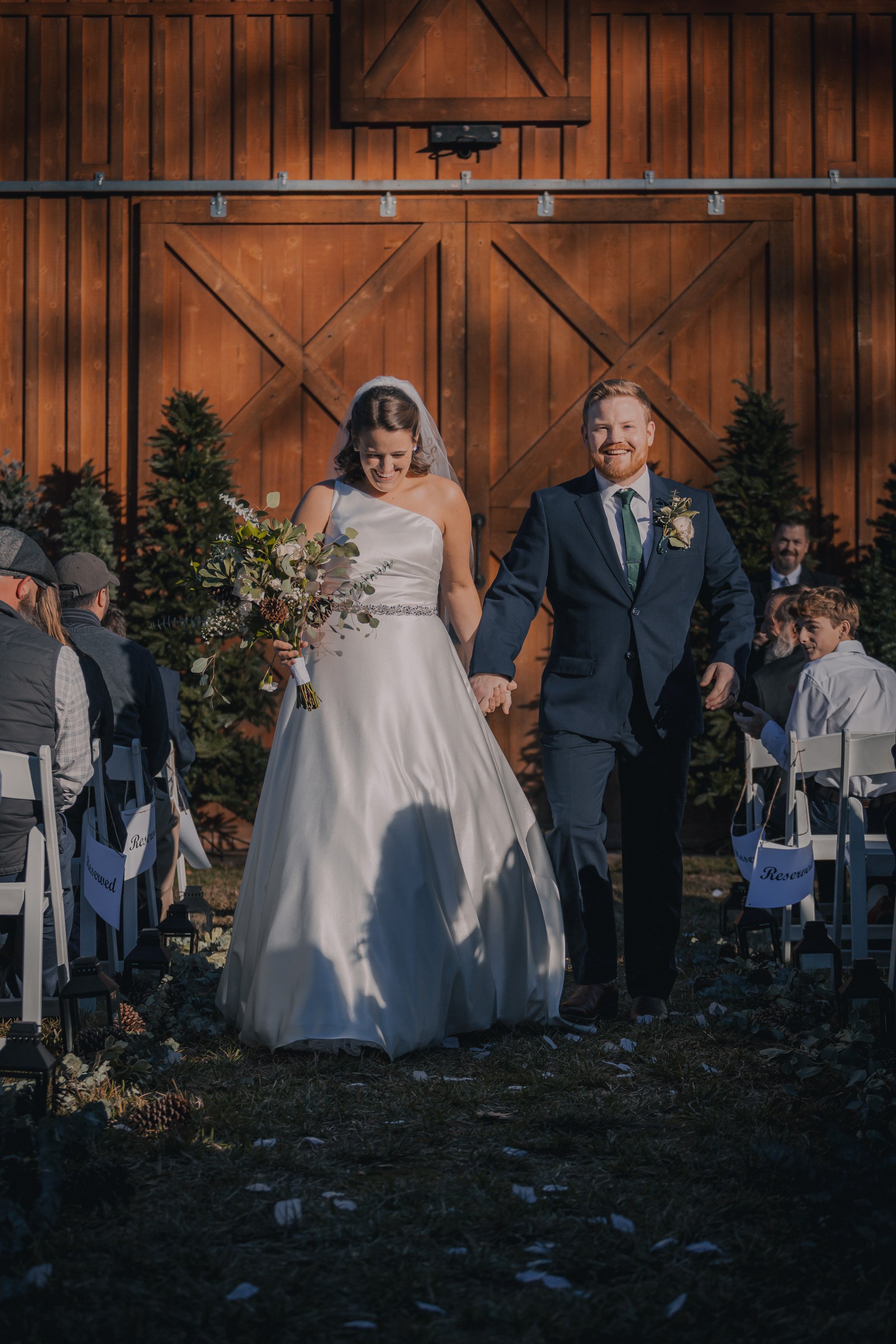 A bride and groom walking hand in hand down an outdoor wedding aisle, smiling, with guests seated on either side, a wooden barn backdrop, and greenery.