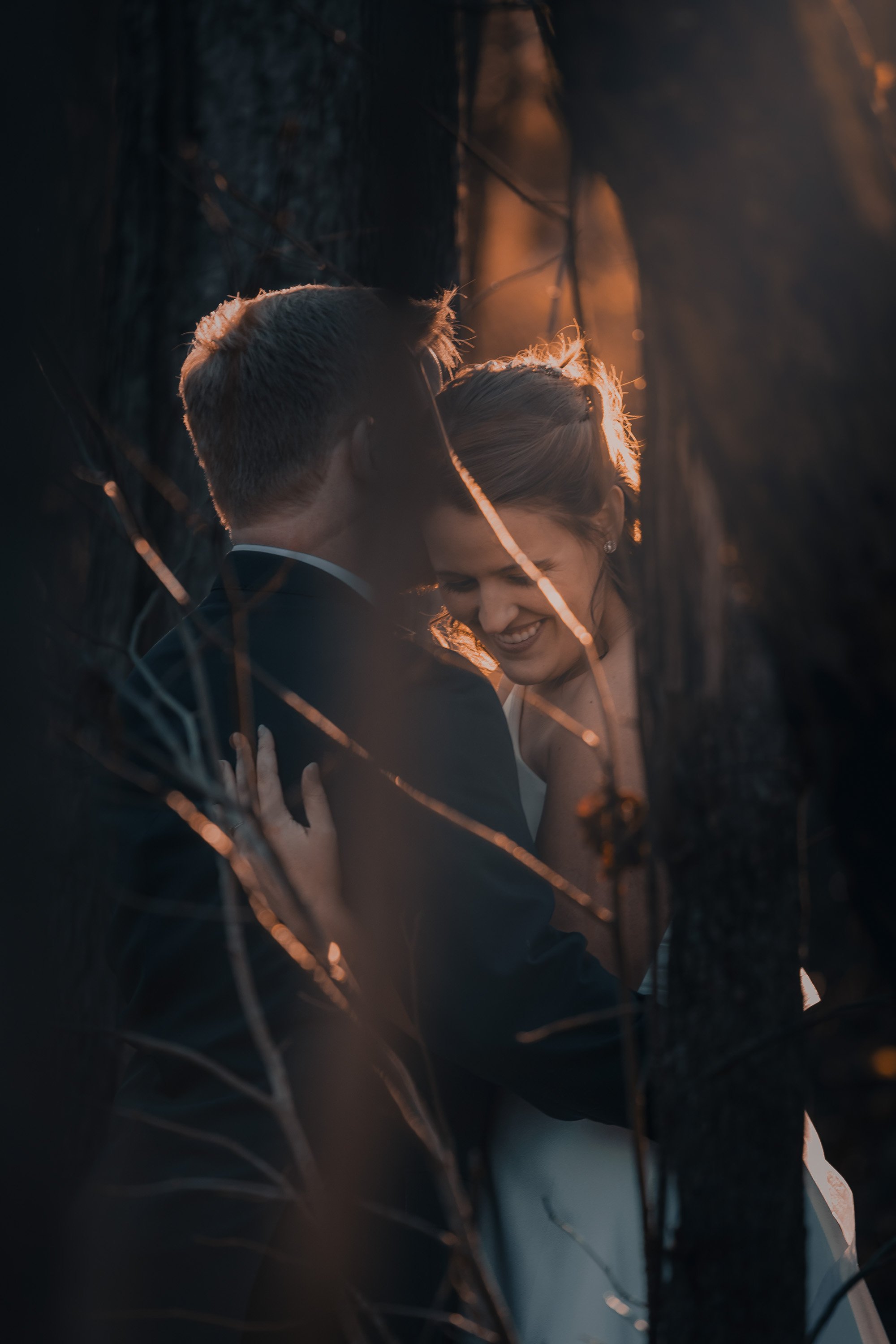 A couple embracing and smiling in a forested area at sunset, with warm sunlight illuminating their faces from behind.