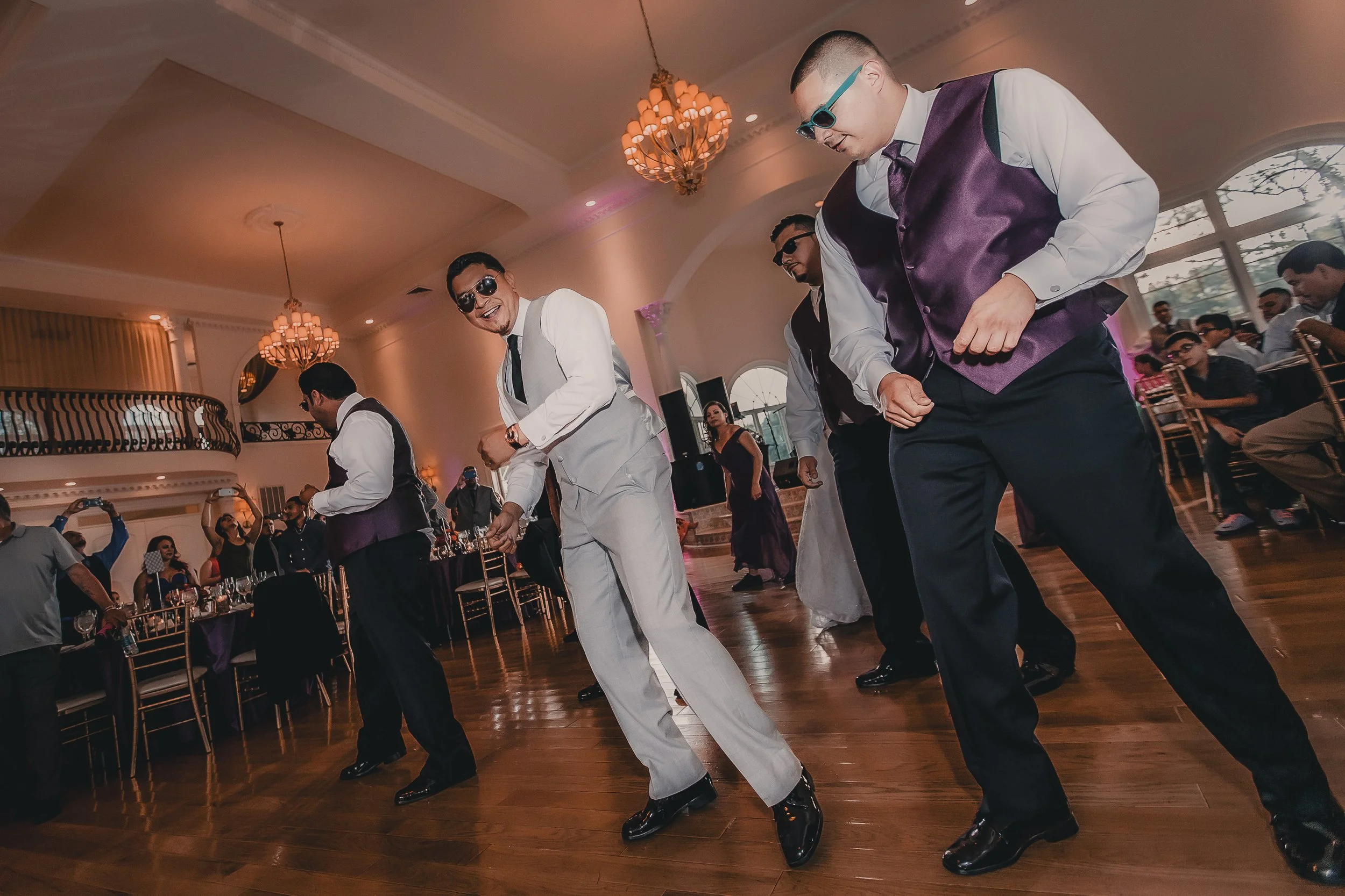 Men dancing at a formal event in a decorated ballroom with chandeliers, seated guests, and a woman in a purple dress in the background.