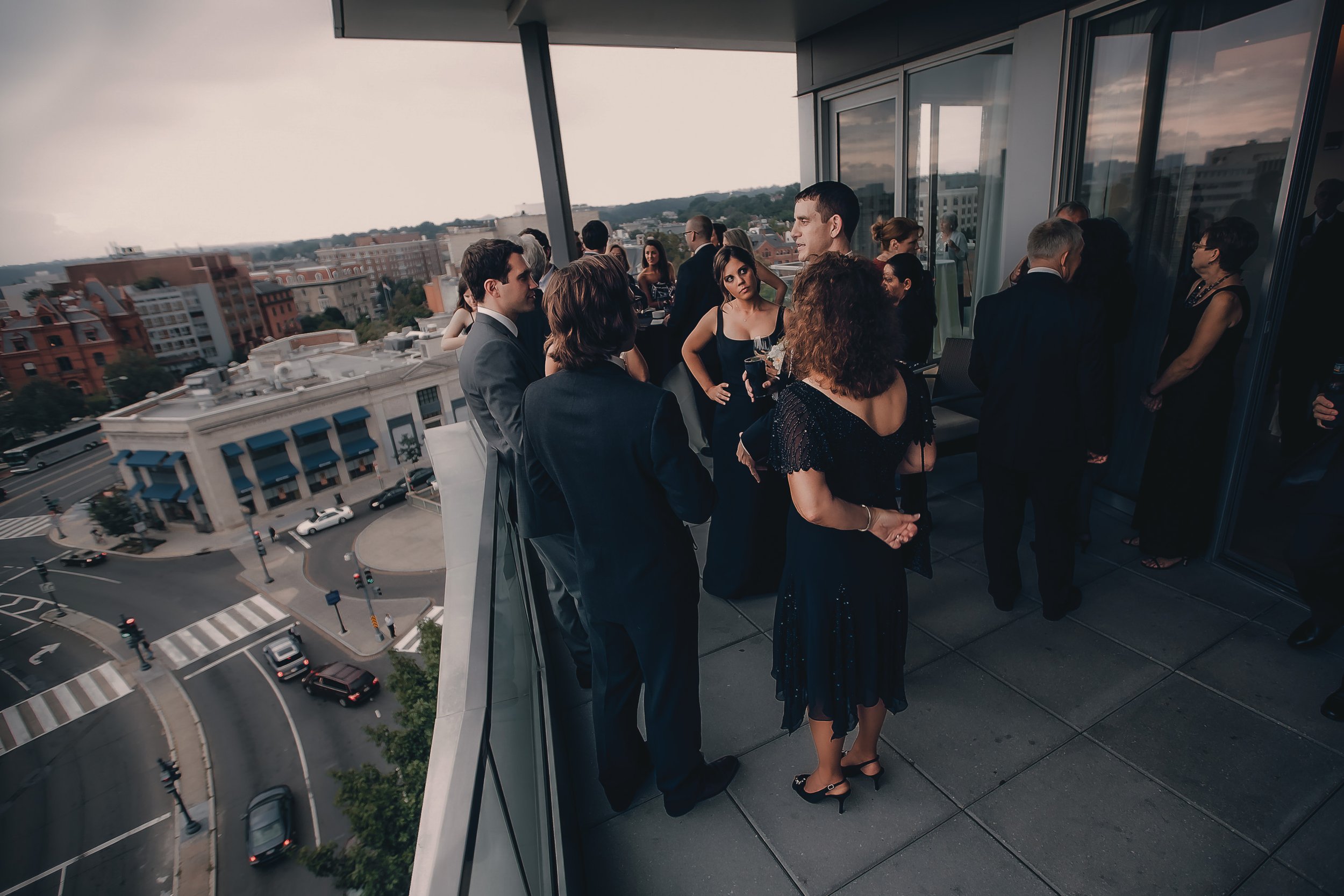 People in formal attire gathered on a city balcony overlooking the streets below during an outdoor event or party in the evening.