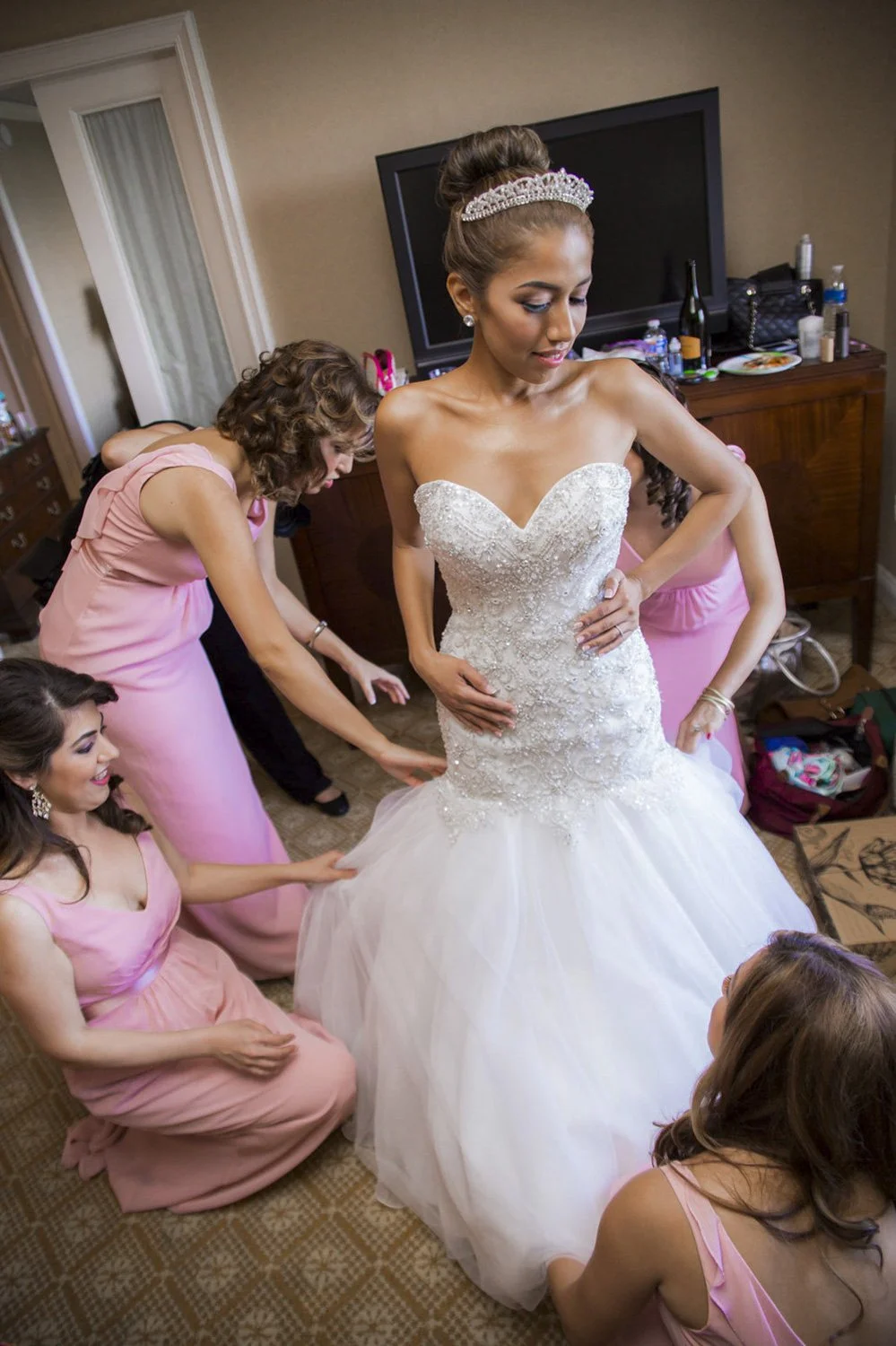 3 bridesmaids help a bride put on her wedding dress in a hotel room.