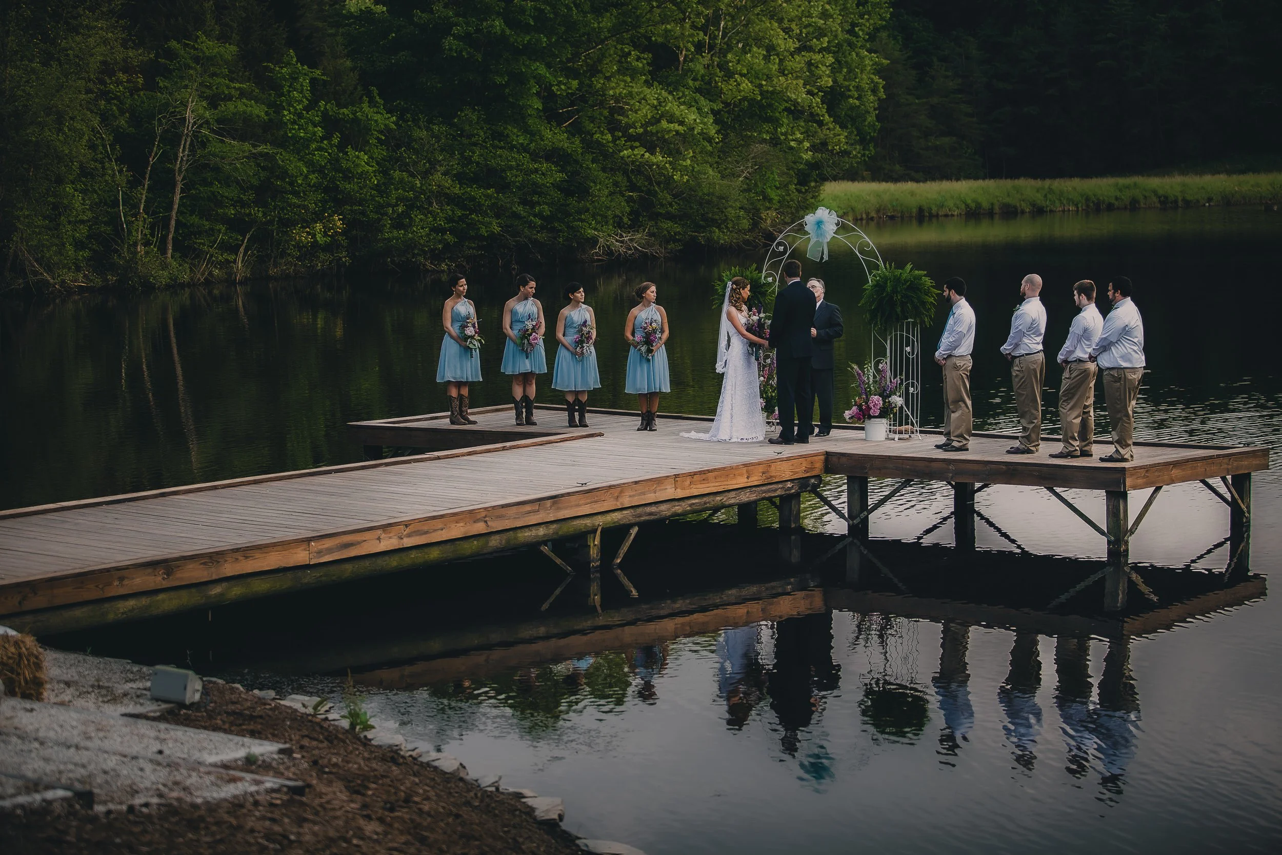 A wedding ceremony taking place on a wooden dock by a lake, with the bride and groom exchanging vows under a white arch decorated with flowers, surrounded by bridesmaids and groomsmen, and trees in the background.