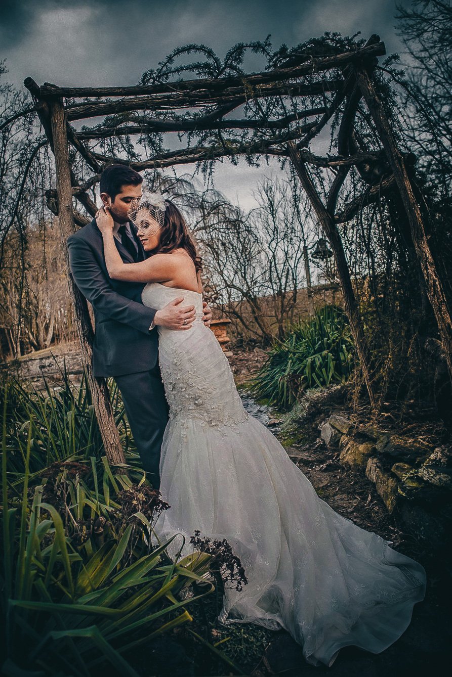 A bride and groom embrace beneath a rustic wooden arch in a forest setting, with overcast skies and bare trees in the background.