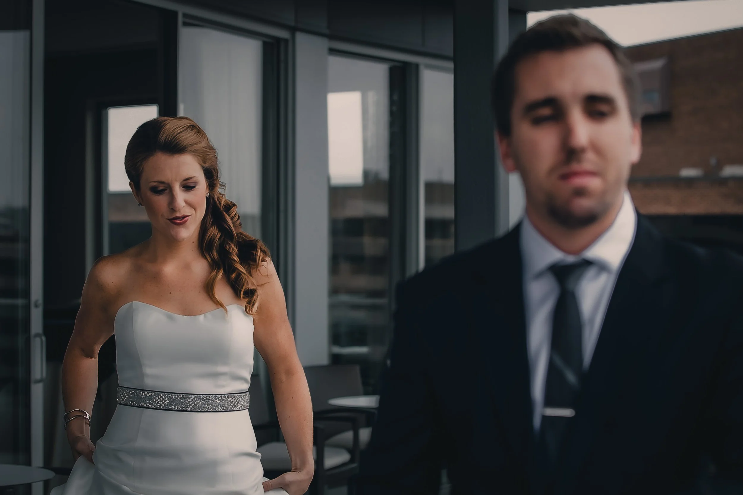 A woman in a white wedding dress with a silver belt and man in a black suit with a tie standing on a balcony with glass doors and city buildings visible in the background.