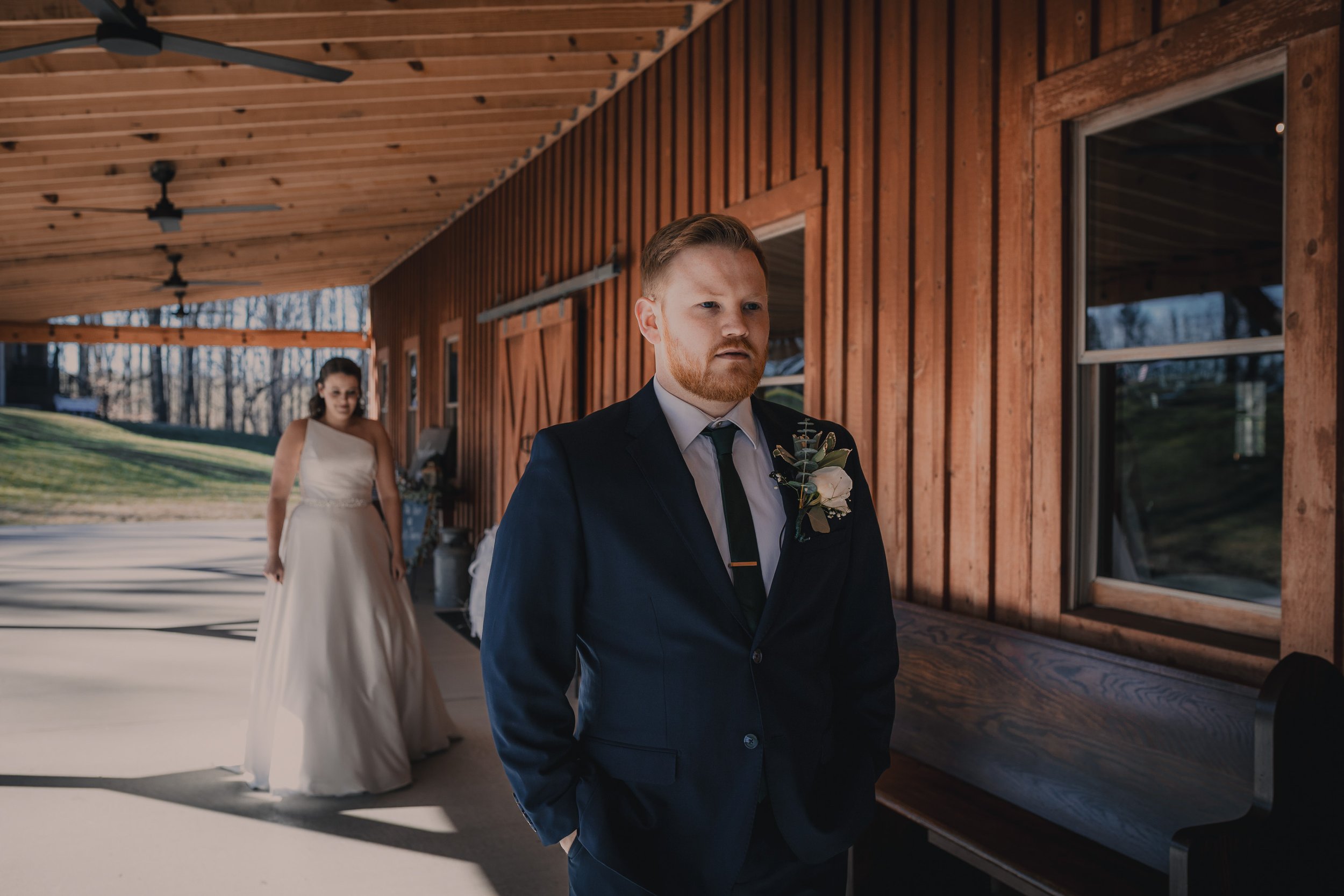 A groom in a navy suit with a boutonniere stands outside a wooden building, looking serious, while a bride in a white wedding dress approaches in the background.