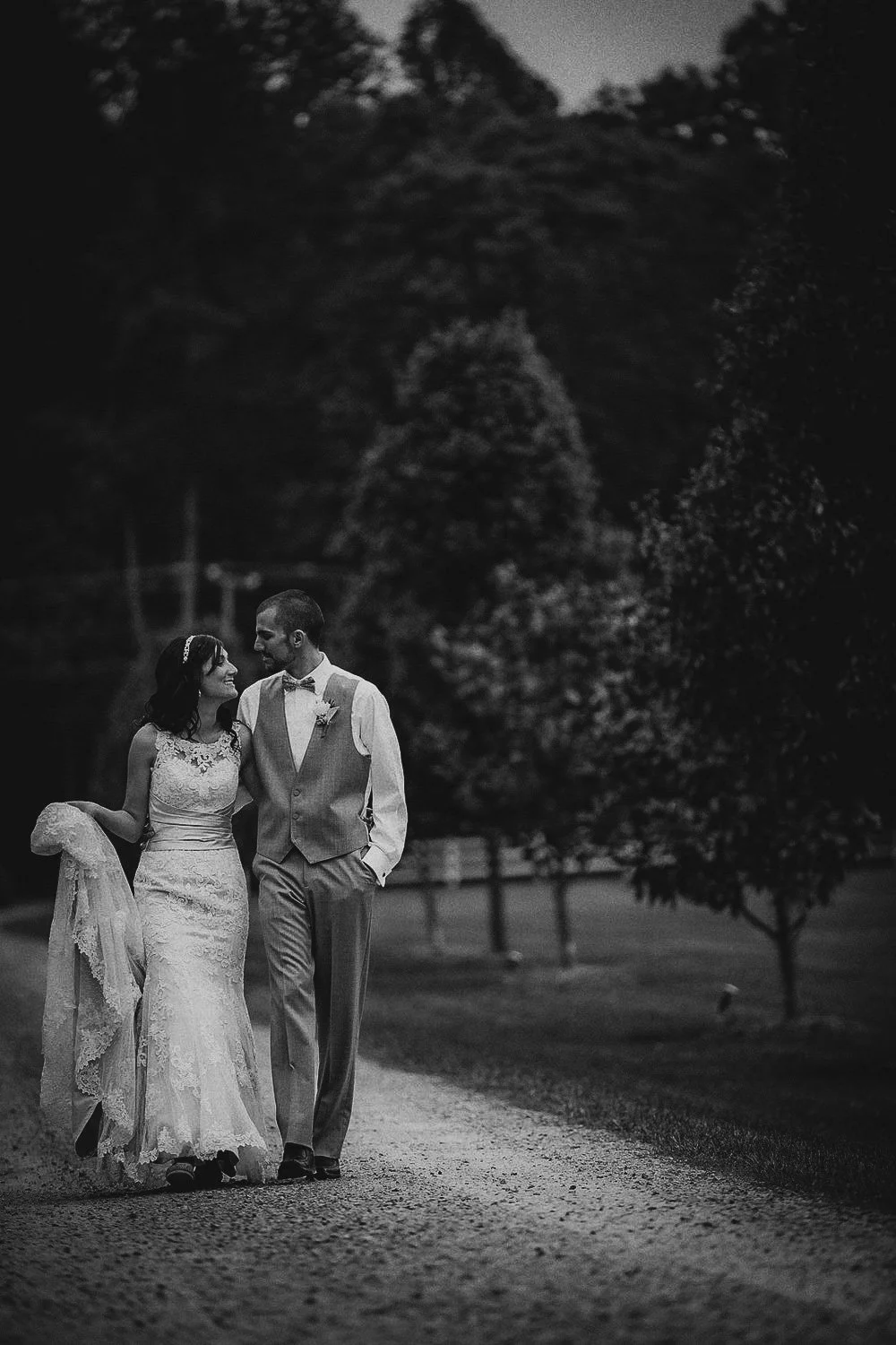 A black and white photo of a newlywed couple walking on a gravel path outdoors at night, surrounded by trees, smiling and looking at each other.