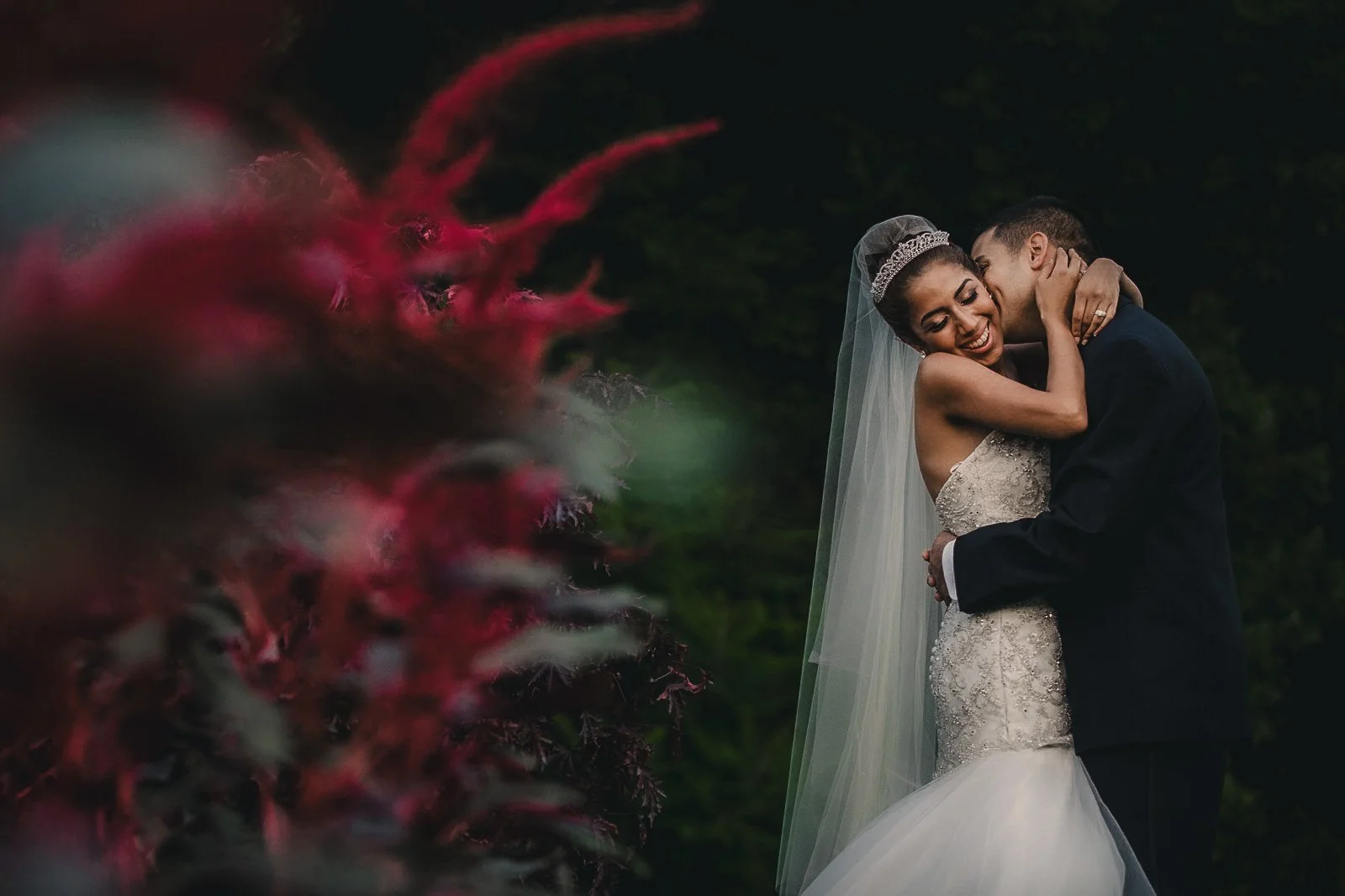 A bride and groom embrace outdoors, the bride smiling with her eyes closed, wearing a white wedding dress and veil, the groom in a dark suit, holding each other closely against a dark background with blurred red flowers in the foreground.