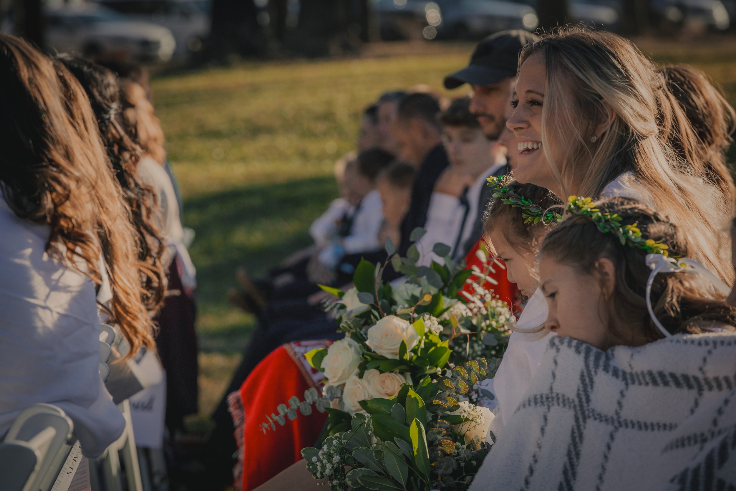 People sitting outdoors, some with floral crowns, with a woman smiling and holding a bouquet of flowers, in a setting with warm sunlight and green grass.