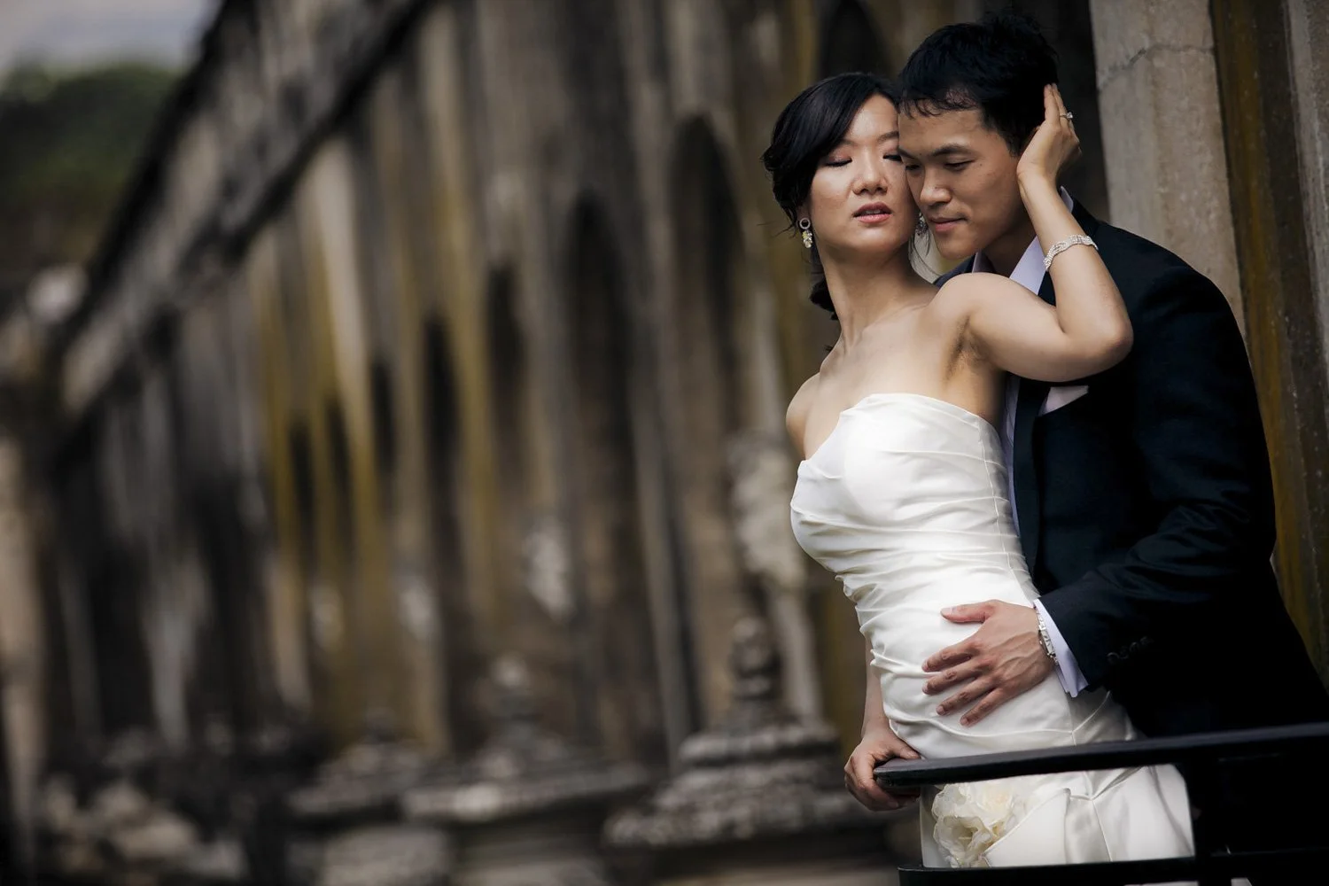 An asian bride and groom embracing against the backdrop of a stone wall with fountain statues.