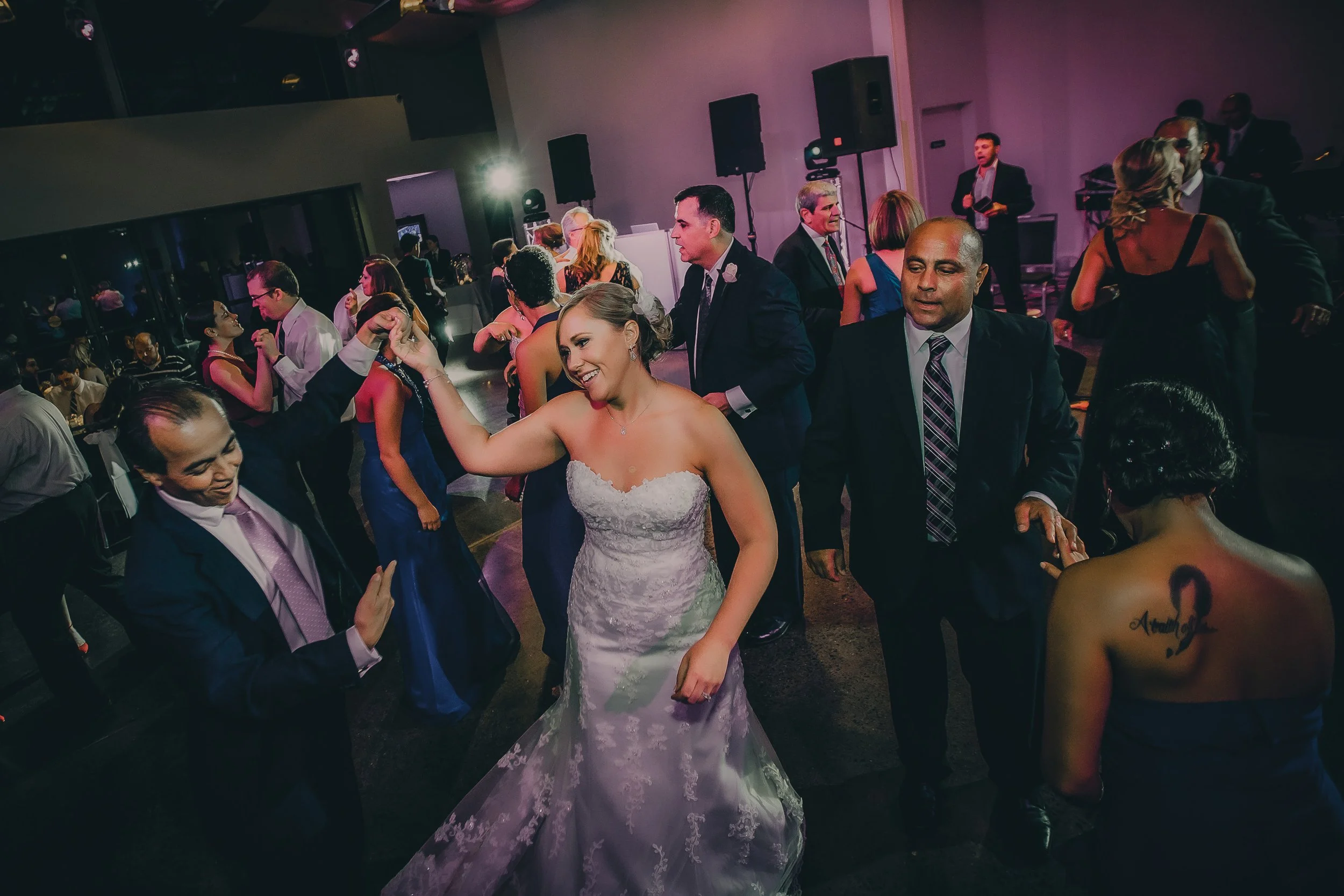 Wedding reception with people dancing, bride in white dress smiling, groom and guests dancing in a dimly lit hall with purple lighting.