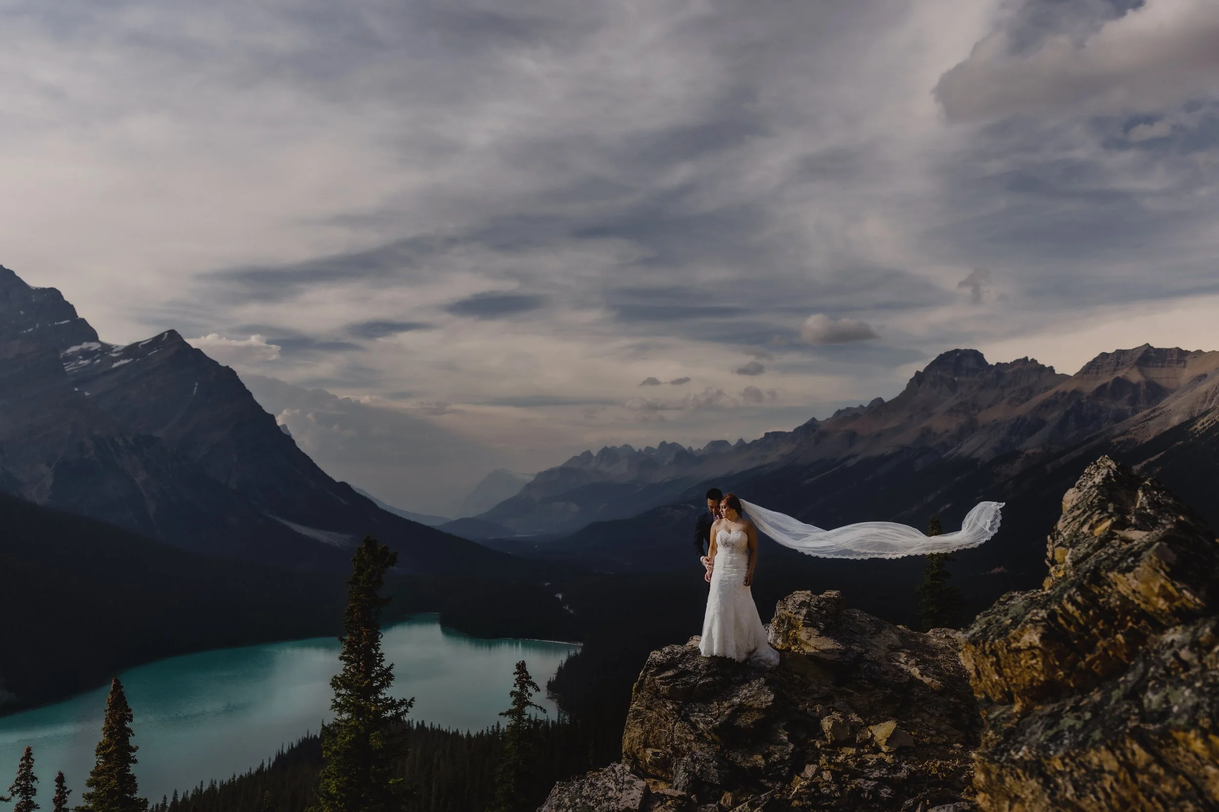 A bride in a white wedding dress and veil standing on a rocky ledge overlooking a lake and mountain range with a cloudy sky.