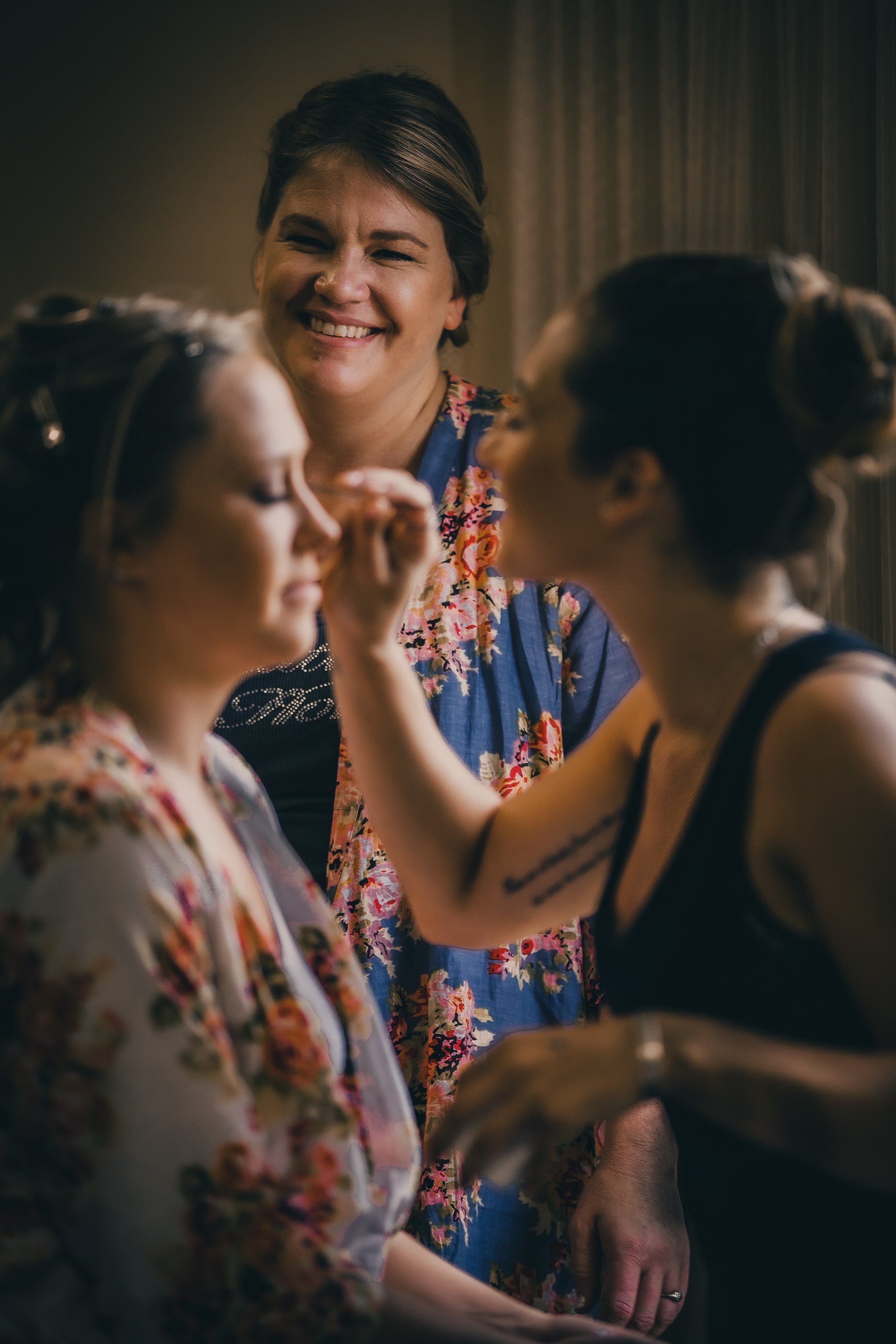 A woman smiling while helping another woman with her makeup, with a third woman observing in the background.