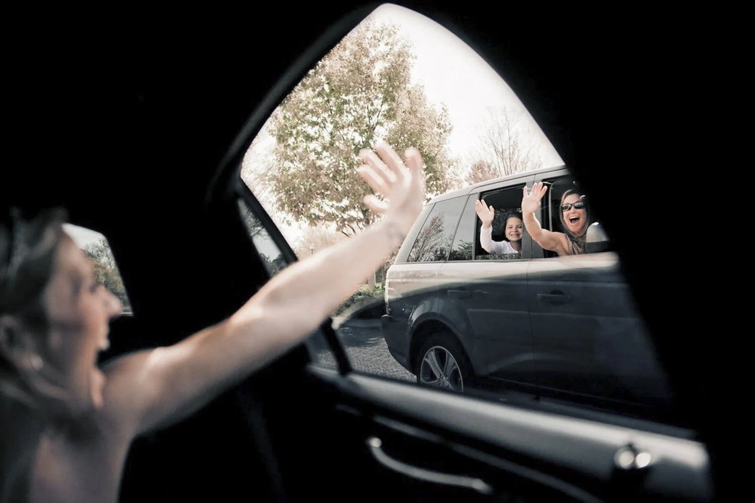 A bride in a limo waving out the window to her family members in the car next to her.