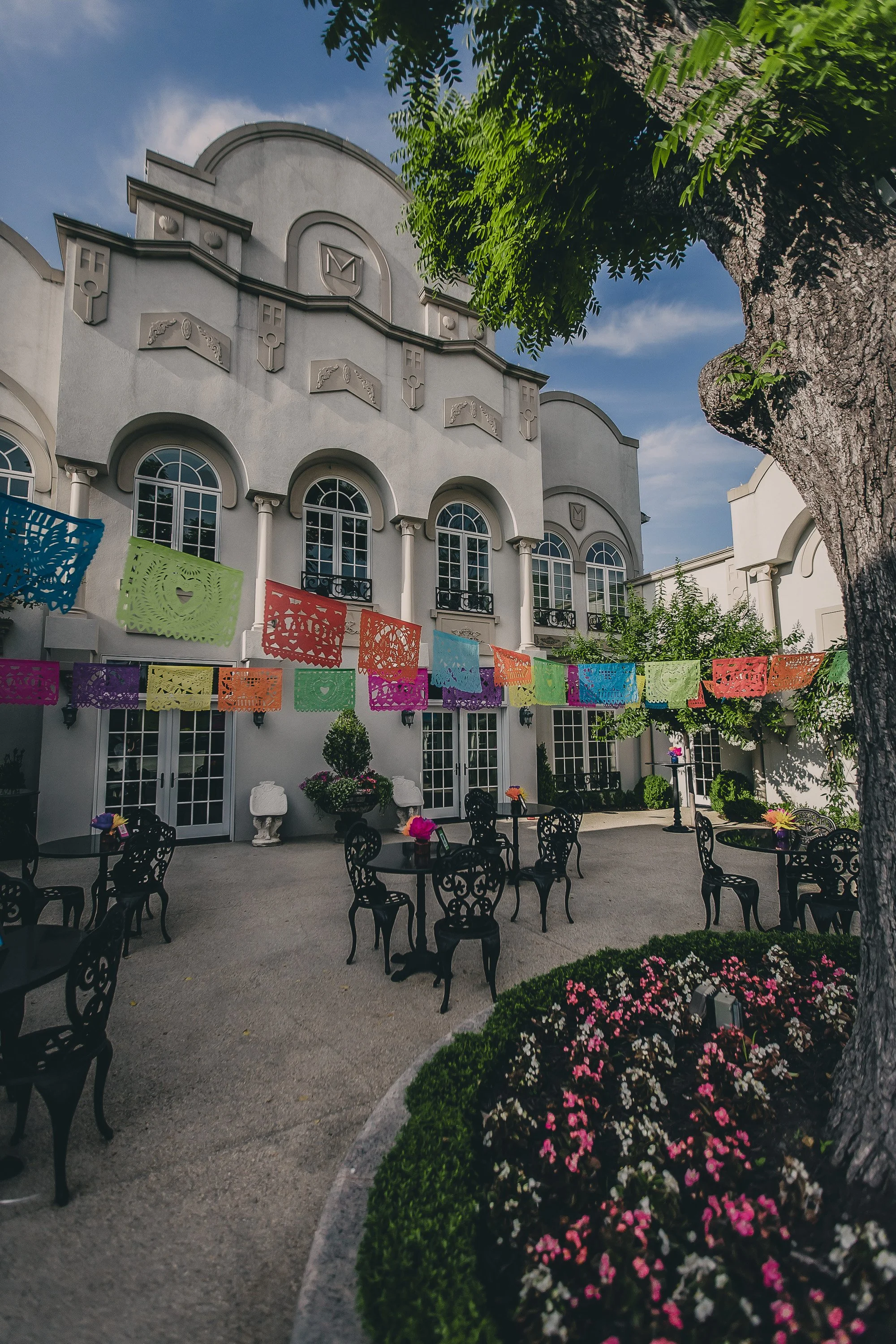 Outdoor patio with black wrought iron chairs and tables, colorful papel picado banners hanging, potted flowers, and a large tree in front of a white building with arched windows.