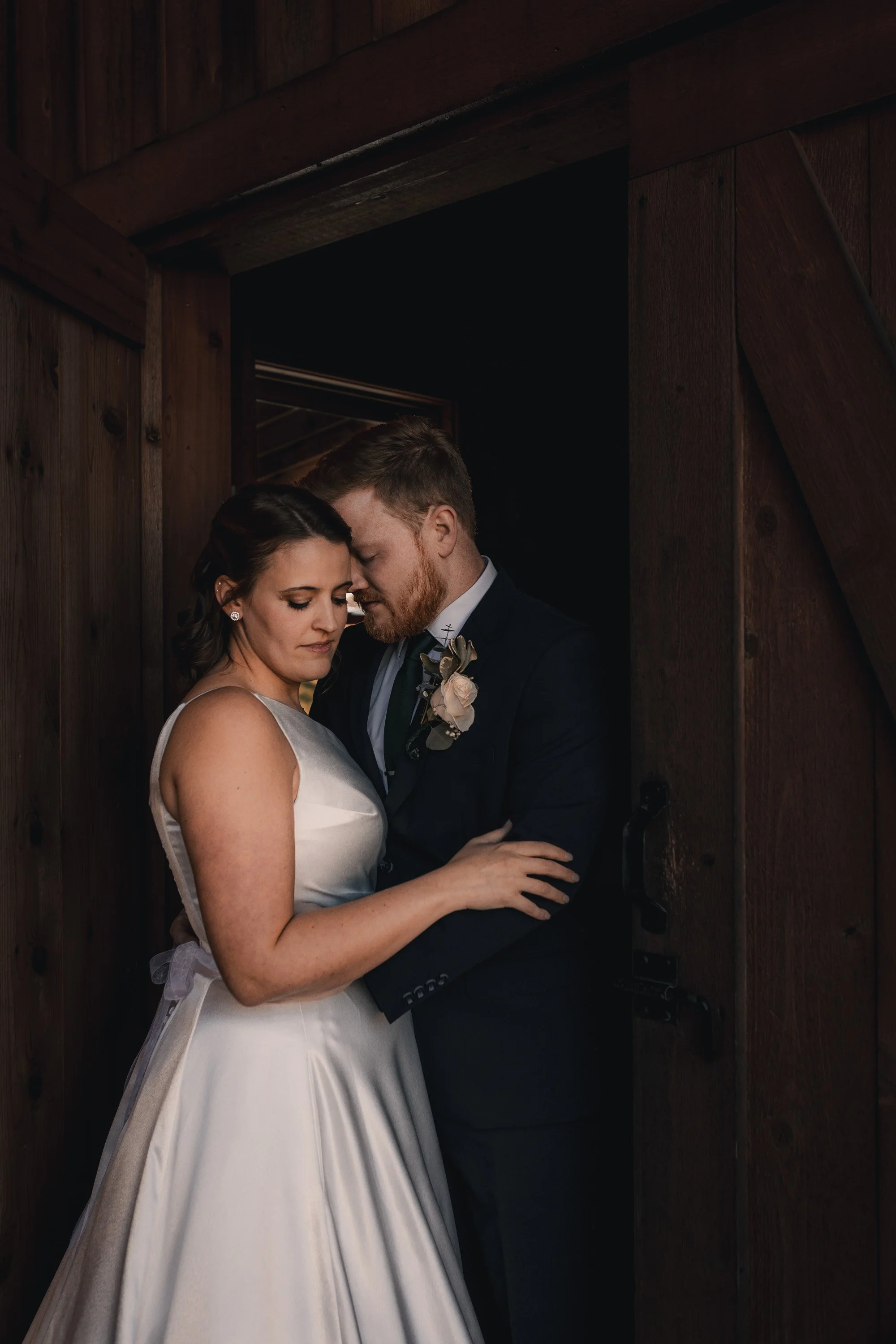 A bride and groom standing close together in a doorway, with their foreheads touching and eyes closed, in a dimly lit wooden setting.