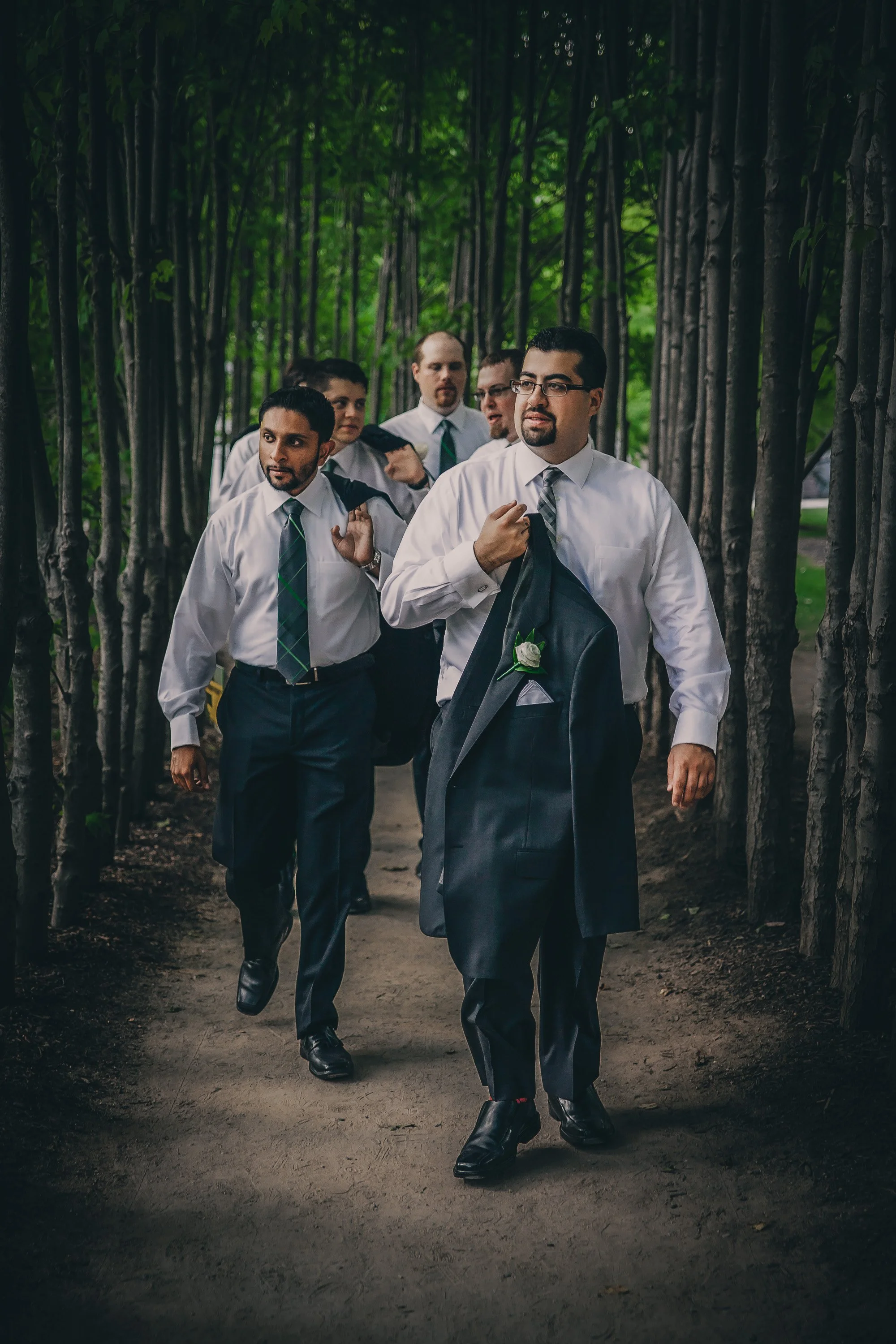 Group of six men dressed in white shirts and ties walking along a wooded pathway, one carrying a suit jacket with a boutonniere.