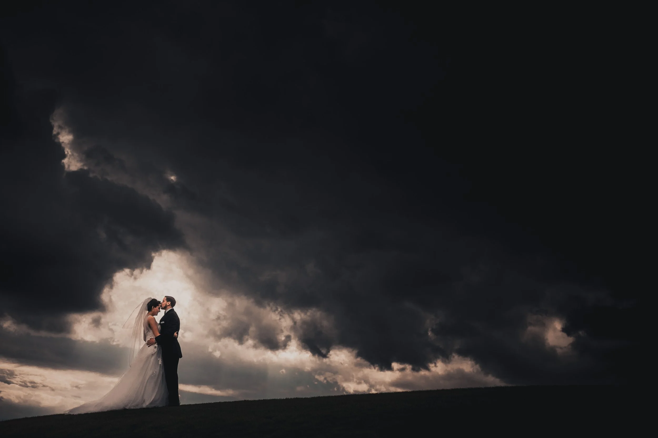 A bride and groom standing close on a hilltop under a dark, stormy sky with heavy clouds.