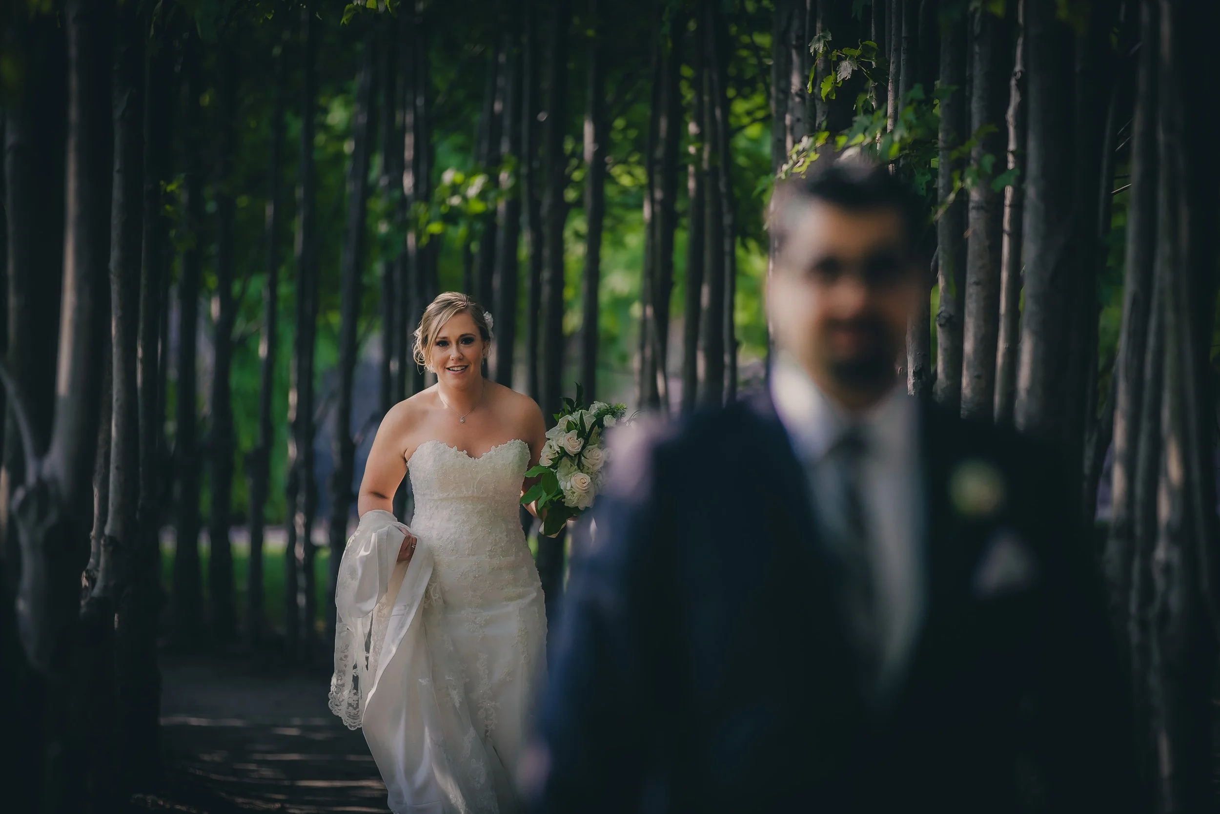 A bride walking through a wooded area, smiling, holding a bouquet, with a groom in the foreground, blurred, dressed in a suit.