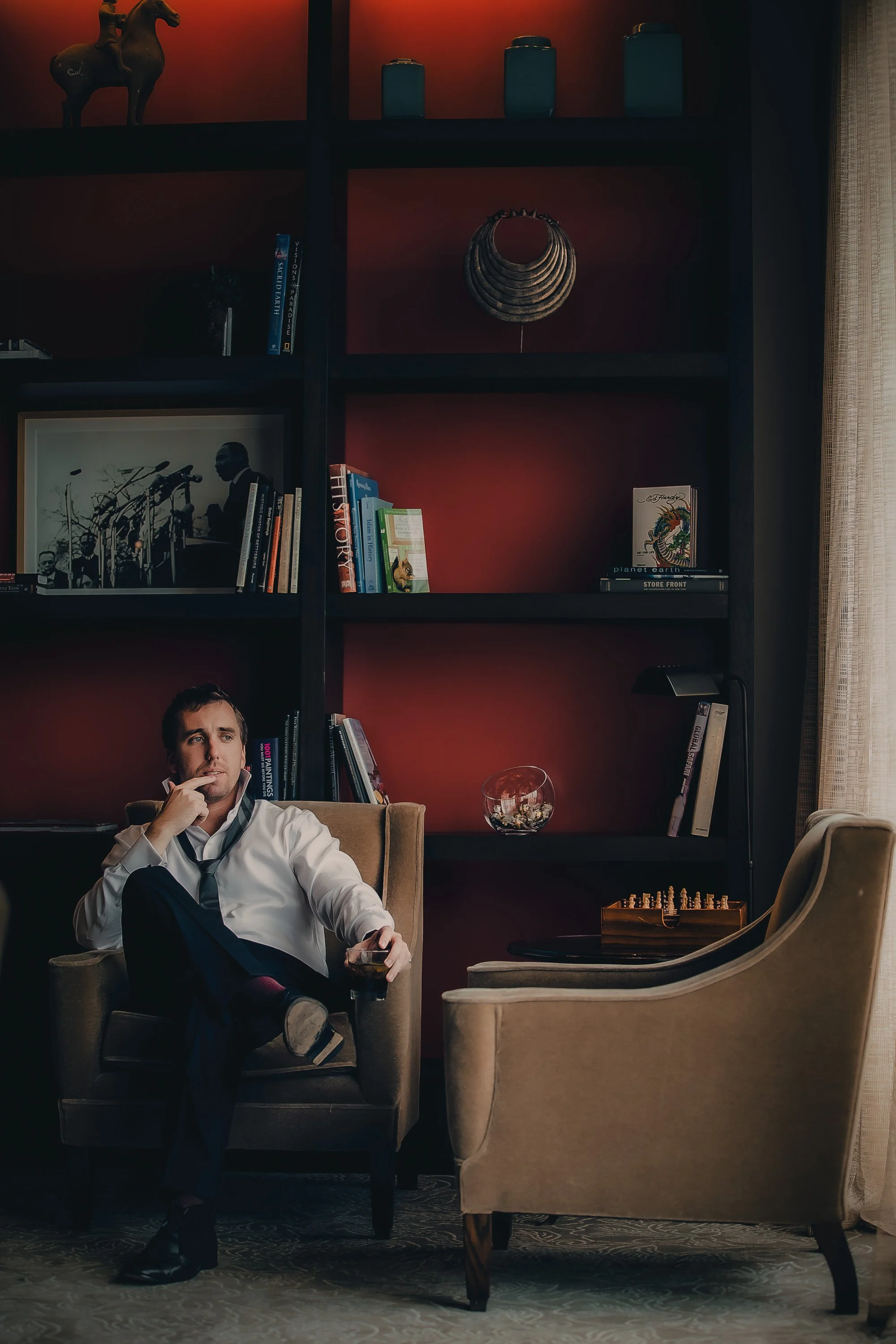 Man sitting in a plush armchair in a dimly lit living room, holding a glass of dark beverage, with a bookshelf filled with books and decorative items behind him.