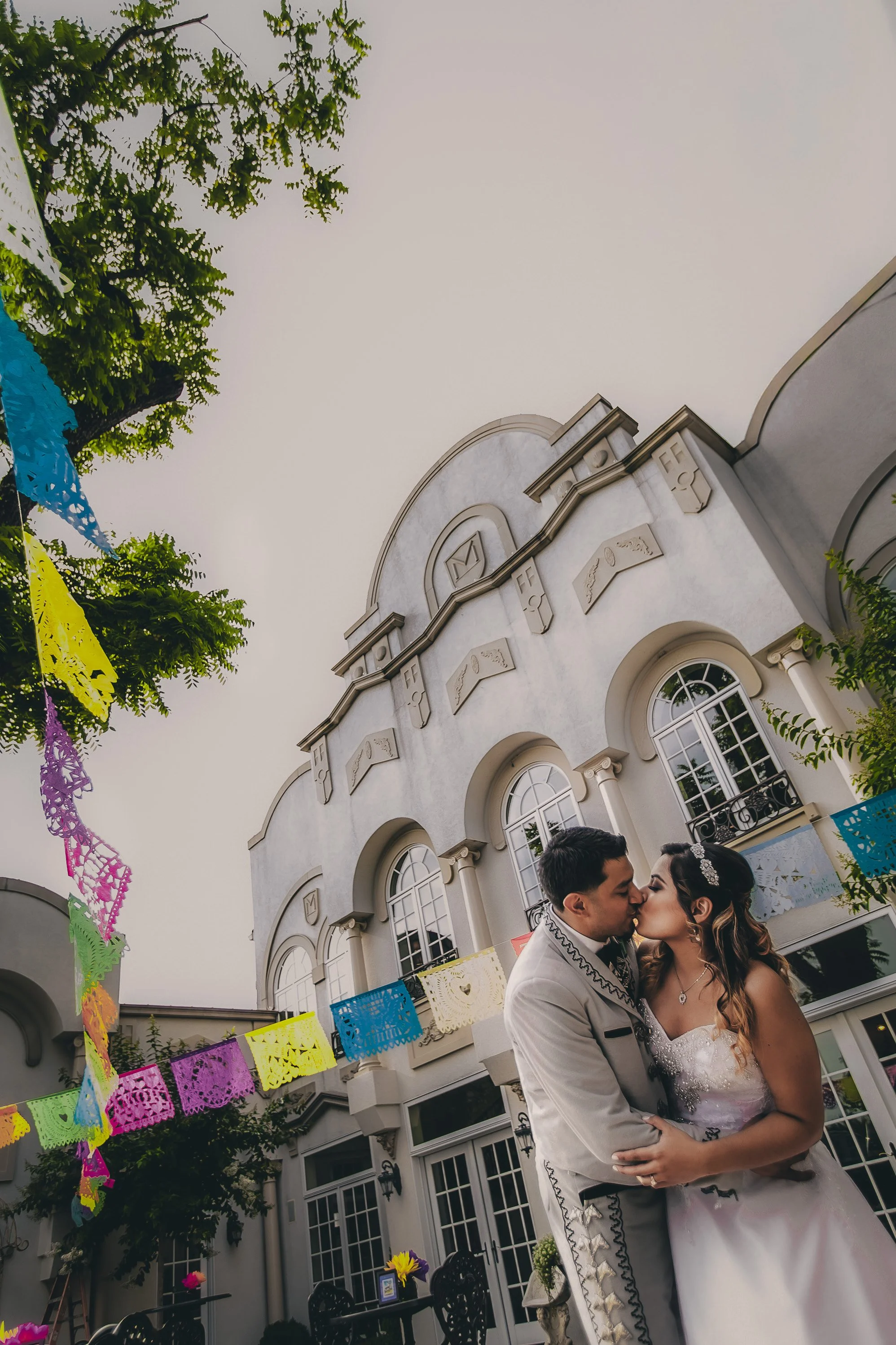 A bride and groom sharing a kiss in front of a white mansion decorated with colorful papel picado banners.