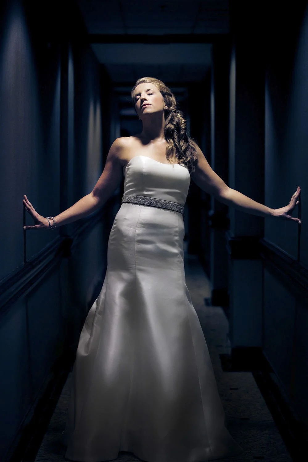 A solo full body portrait of a bride posing in a dark hallway, lifting her face to be illuminated by an overhead light.