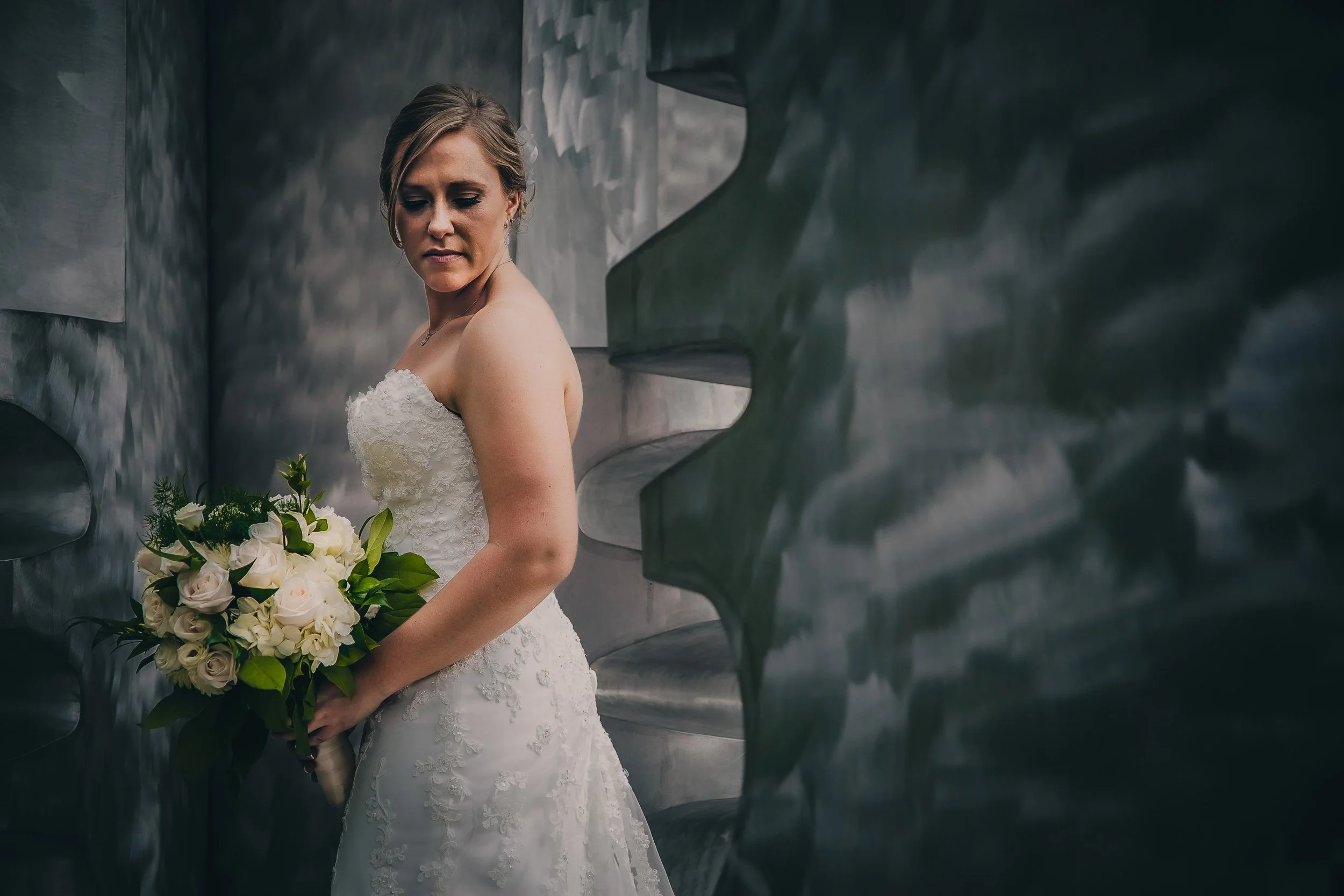 A bride in a white wedding dress holding a bouquet of white roses and greenery, standing against a metallic, textured wall, looking down with a contemplative expression.
