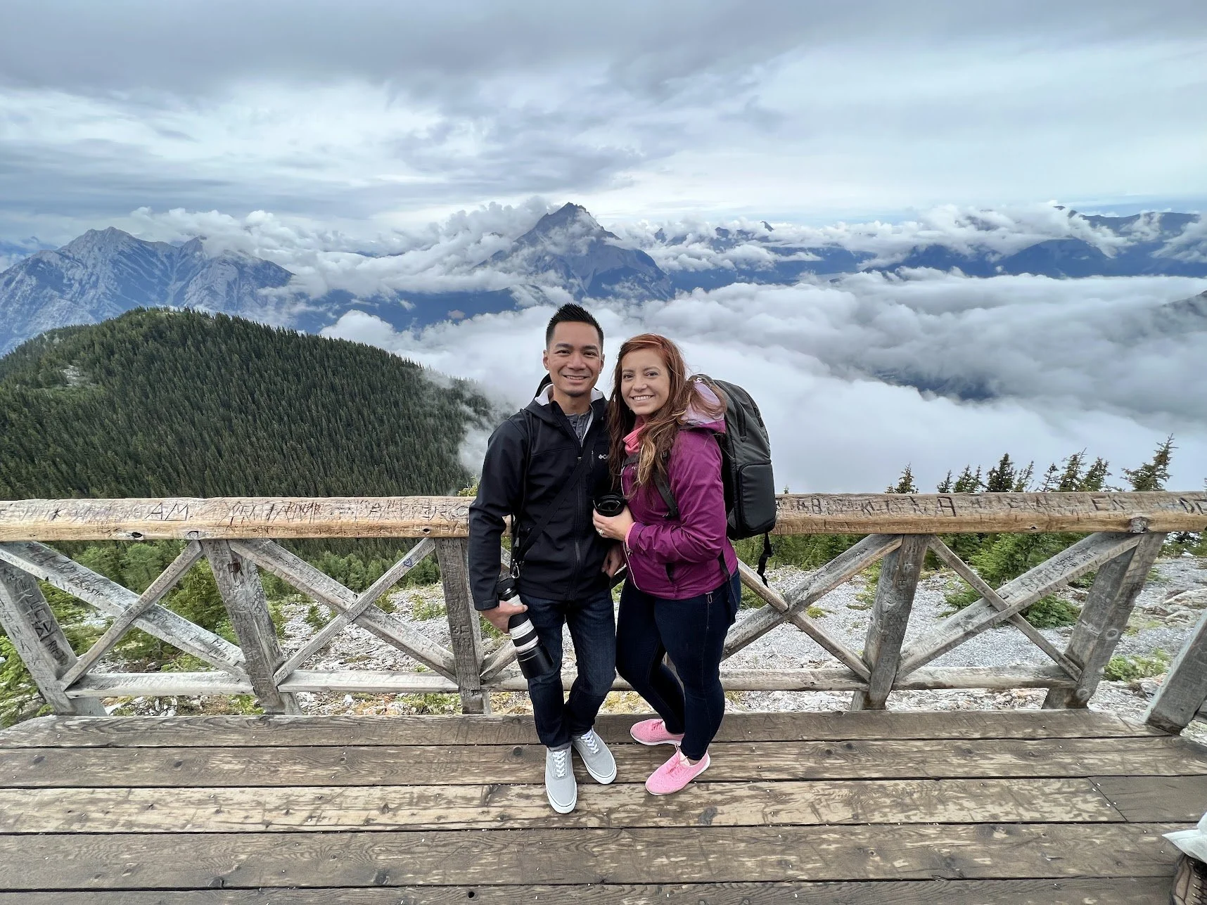 A smiling man and woman with backpacks and cameras standing on a wooden platform in the mountains, surrounded by clouds and lush green trees.