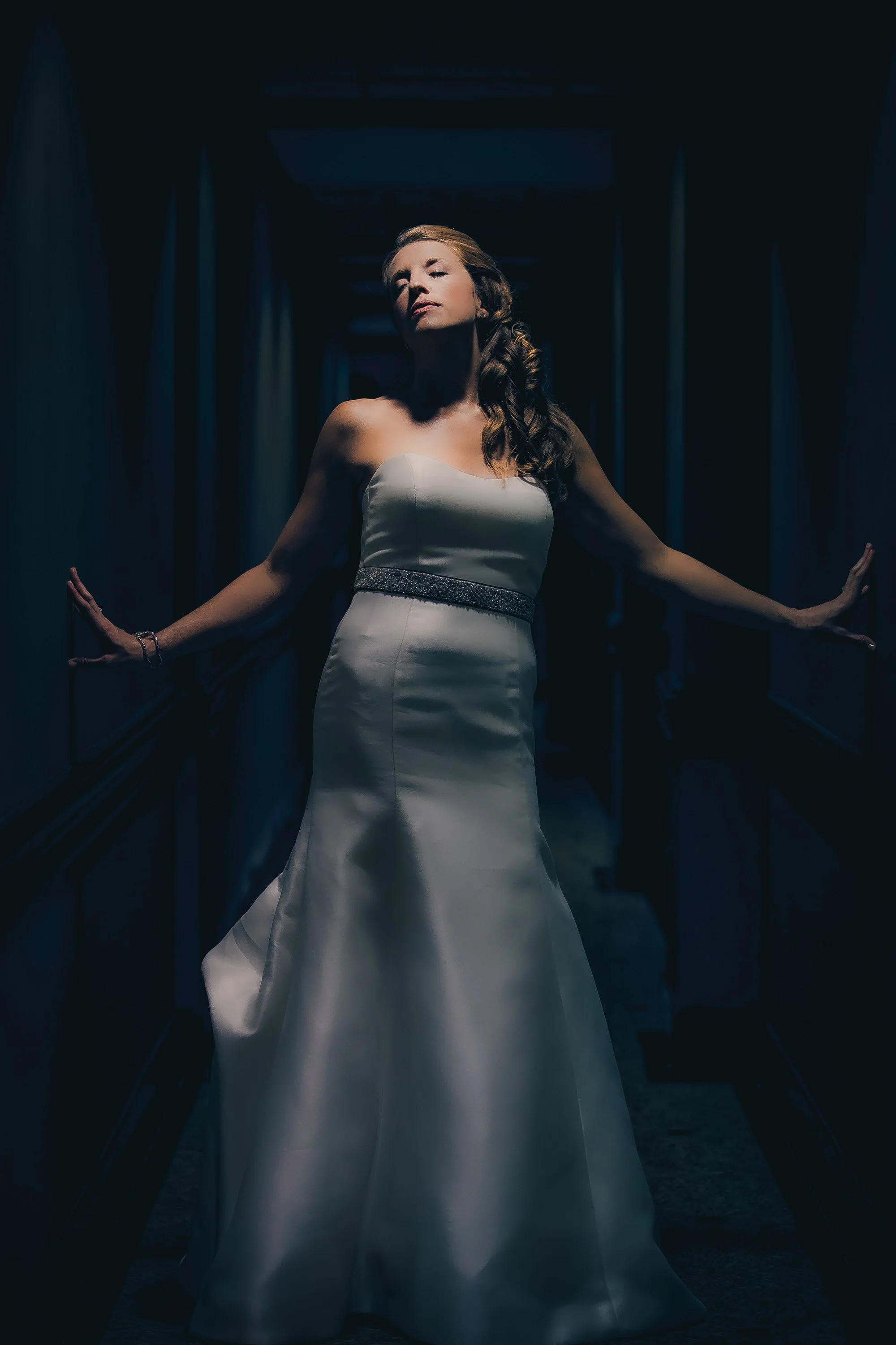 A woman in a strapless white gown standing in a dark hallway with her arms outstretched, looking confidently ahead.