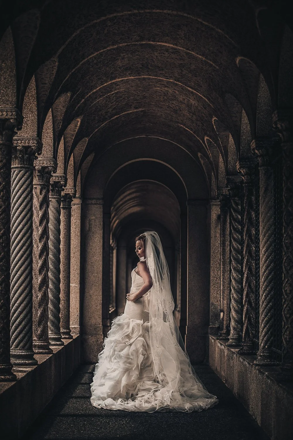 A bride in a strapless wedding dress with a long veil stands in an arched stone corridor, looking down with one hand on her hip.