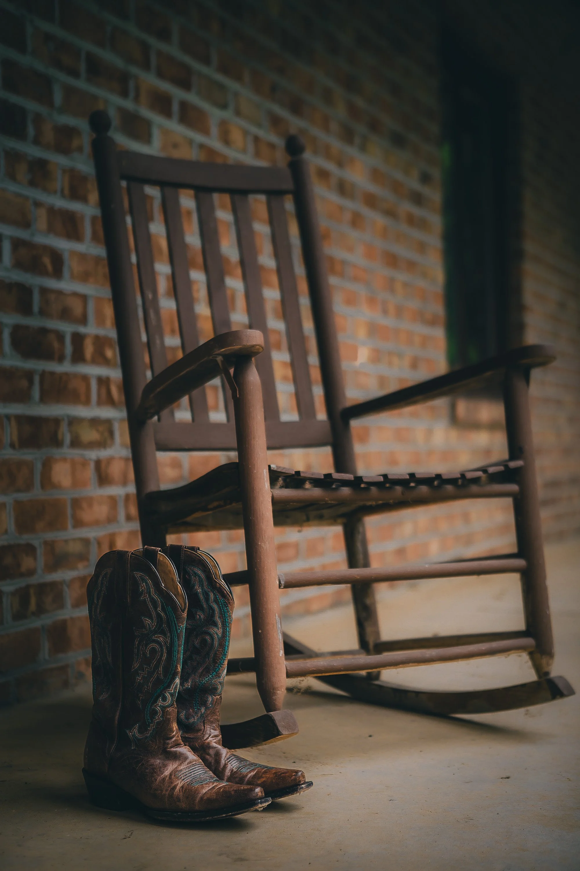 A pair of cowboy boots in front of an old wooden rocking chair against a brick wall.