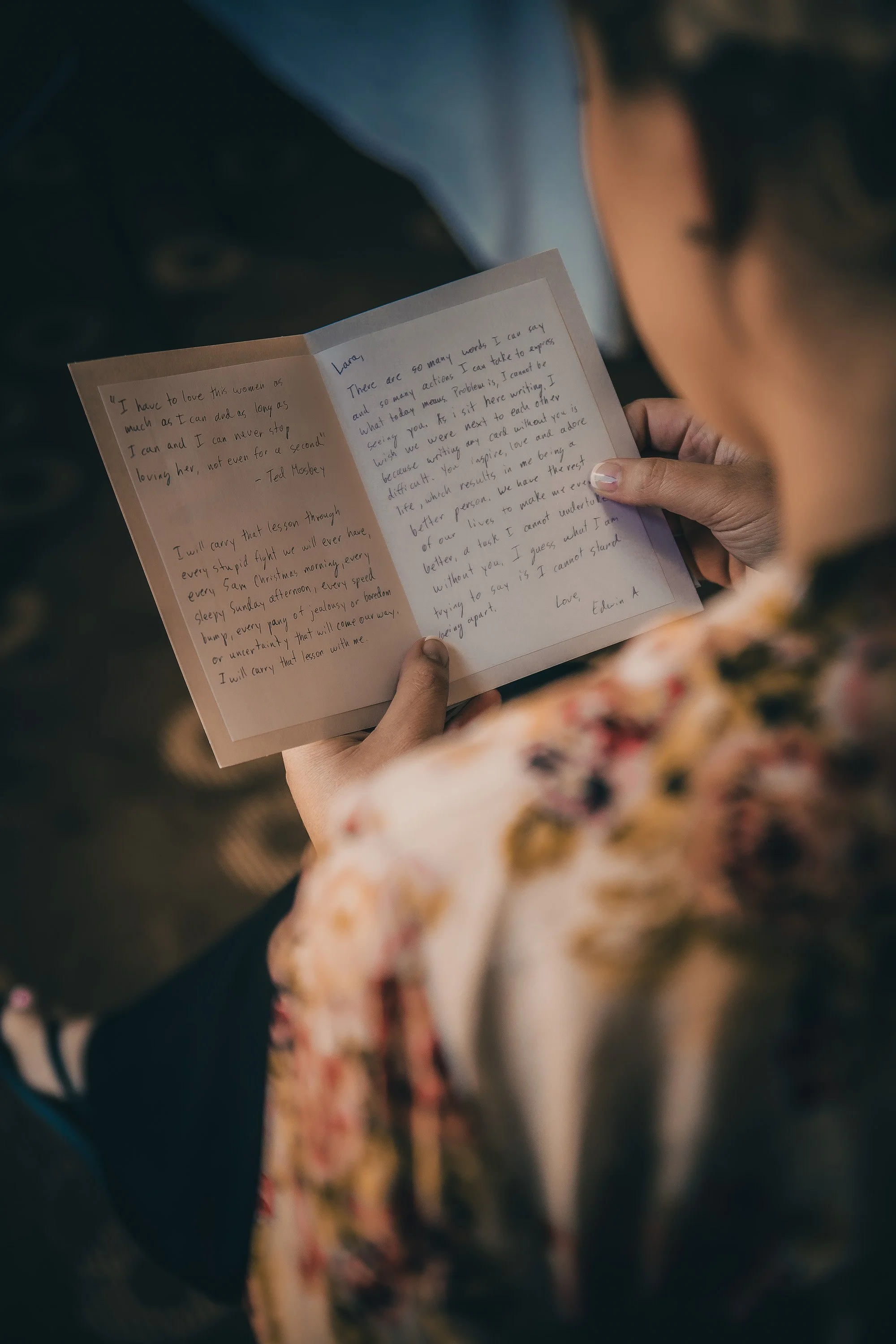 Person reading a handwritten letter inside a paper card, with a floral-patterned dress and denim jacket visible. The person's face is not shown.