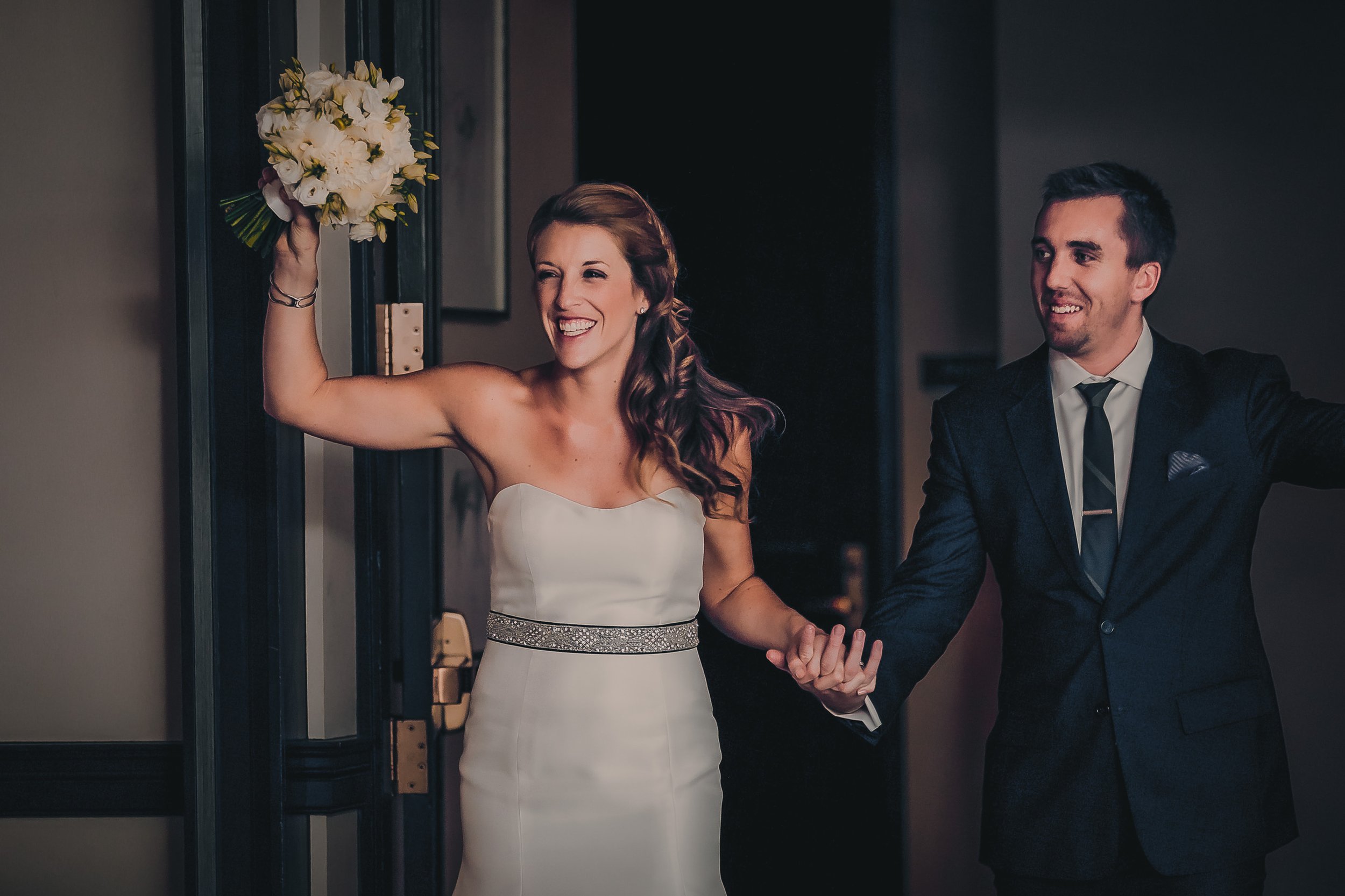 A smiling woman in a white wedding dress holding a bouquet of flowers and a man in a suit holding her hand at a wedding or celebration event.