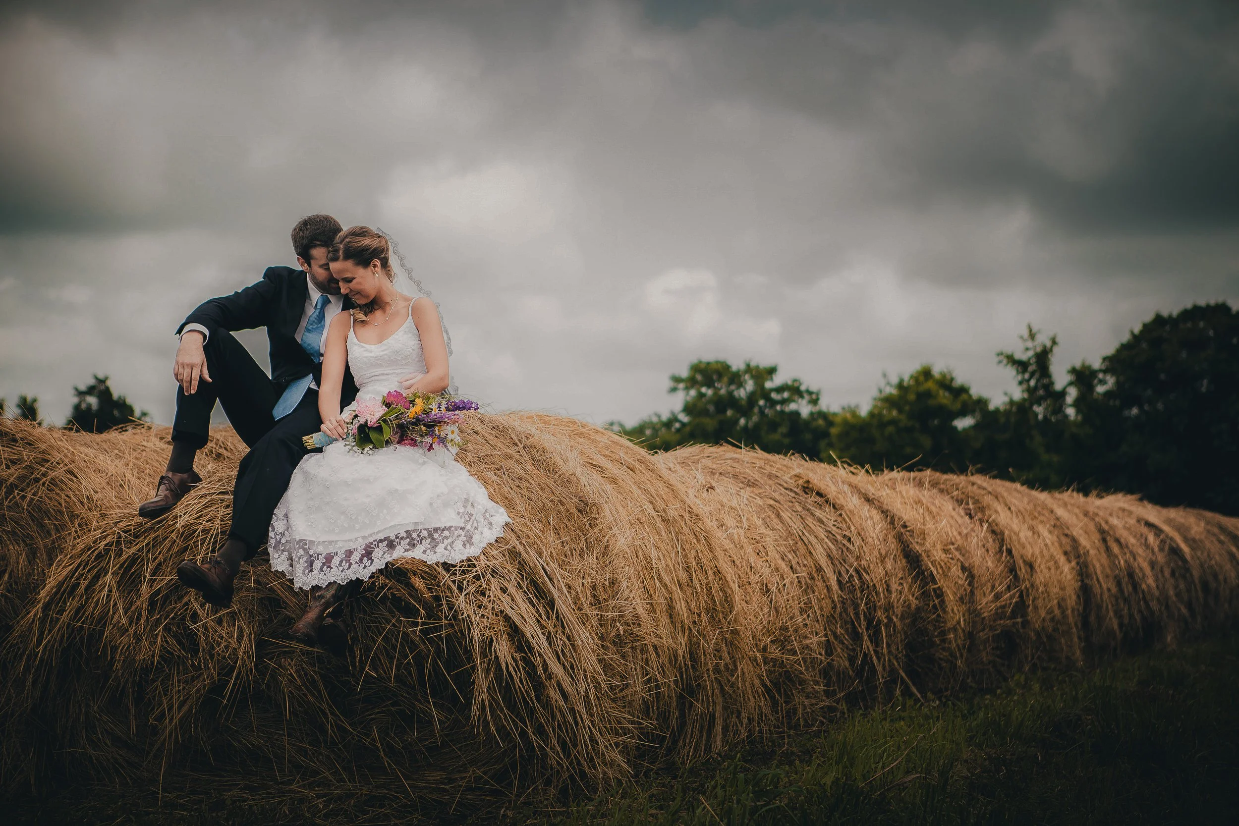 A bride and groom sitting on a hay bale in a field during a cloudy day, with the bride holding a bouquet of flowers.