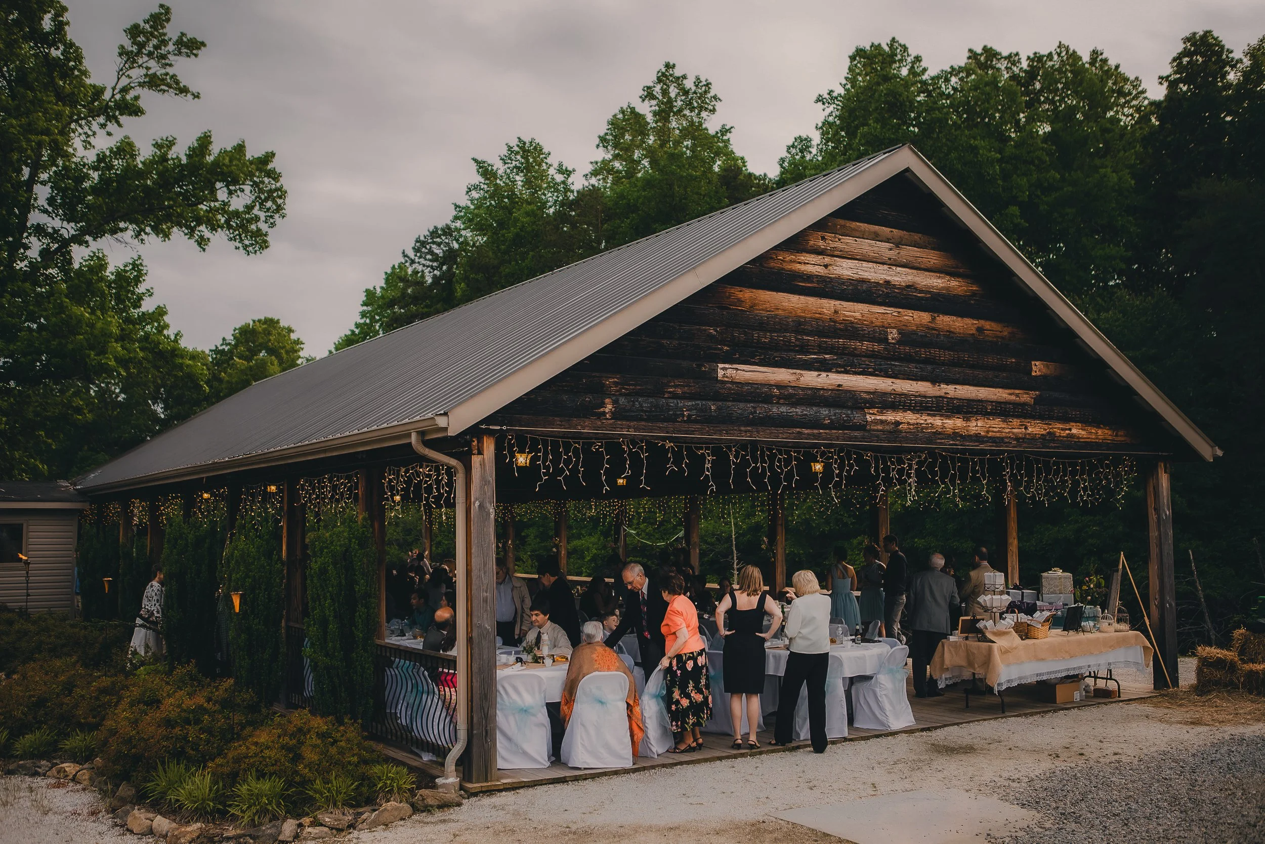 People gathered under a rustic wooden pavilion decorated with string lights, attending a social event or celebration outdoors surrounded by trees.
