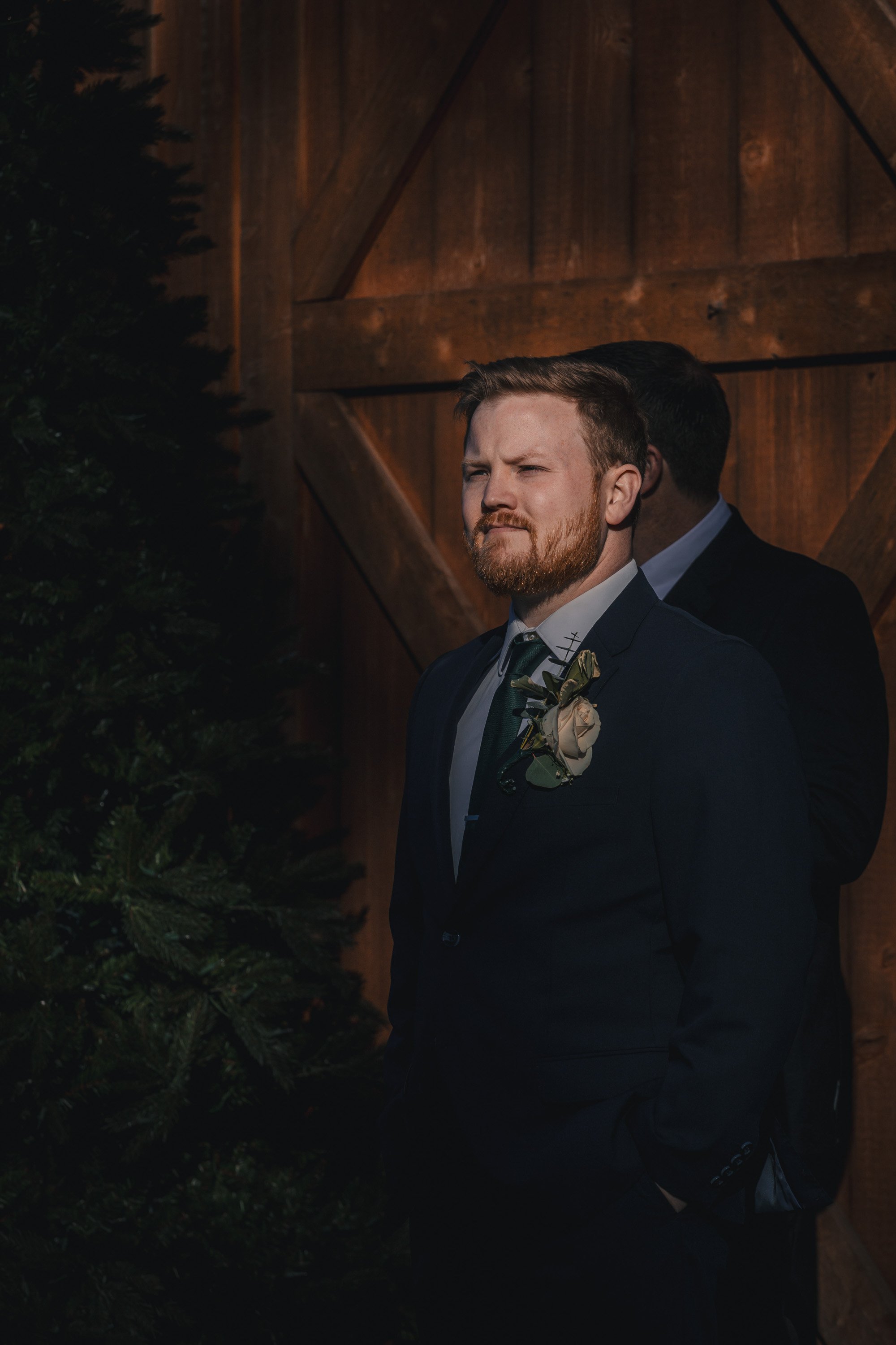 A man with a beard and red hair, dressed in a dark suit with a boutonniere, stands with his hands in his pockets in front of a wooden wall, with a partial view of another person behind him.